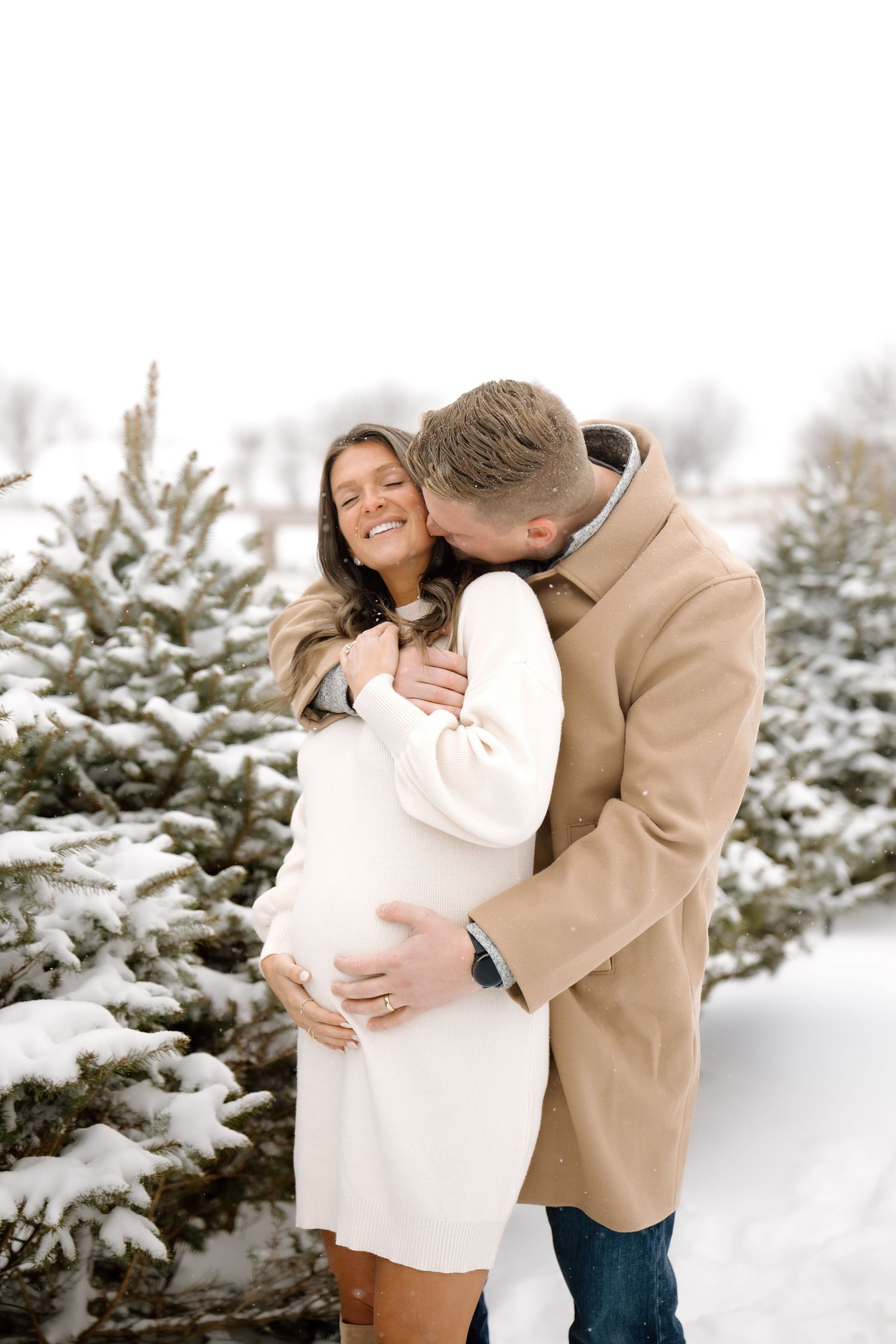 Man in tan coat kisses pregnant partner in white dress among snow-covered evergreen trees.