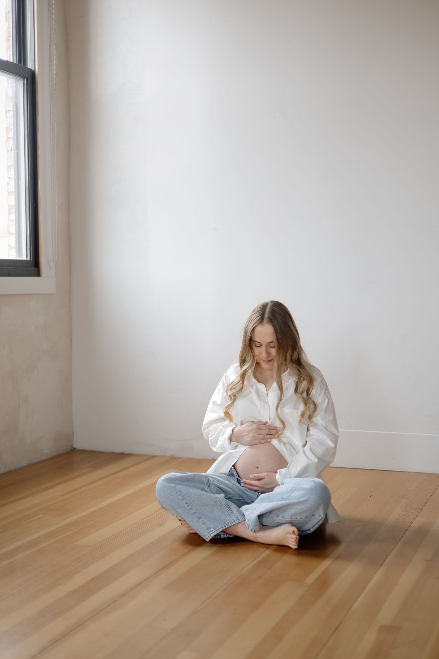 A pregnant woman in a white shirt and jeans sits cross-legged on a wooden floor holding her bump.
