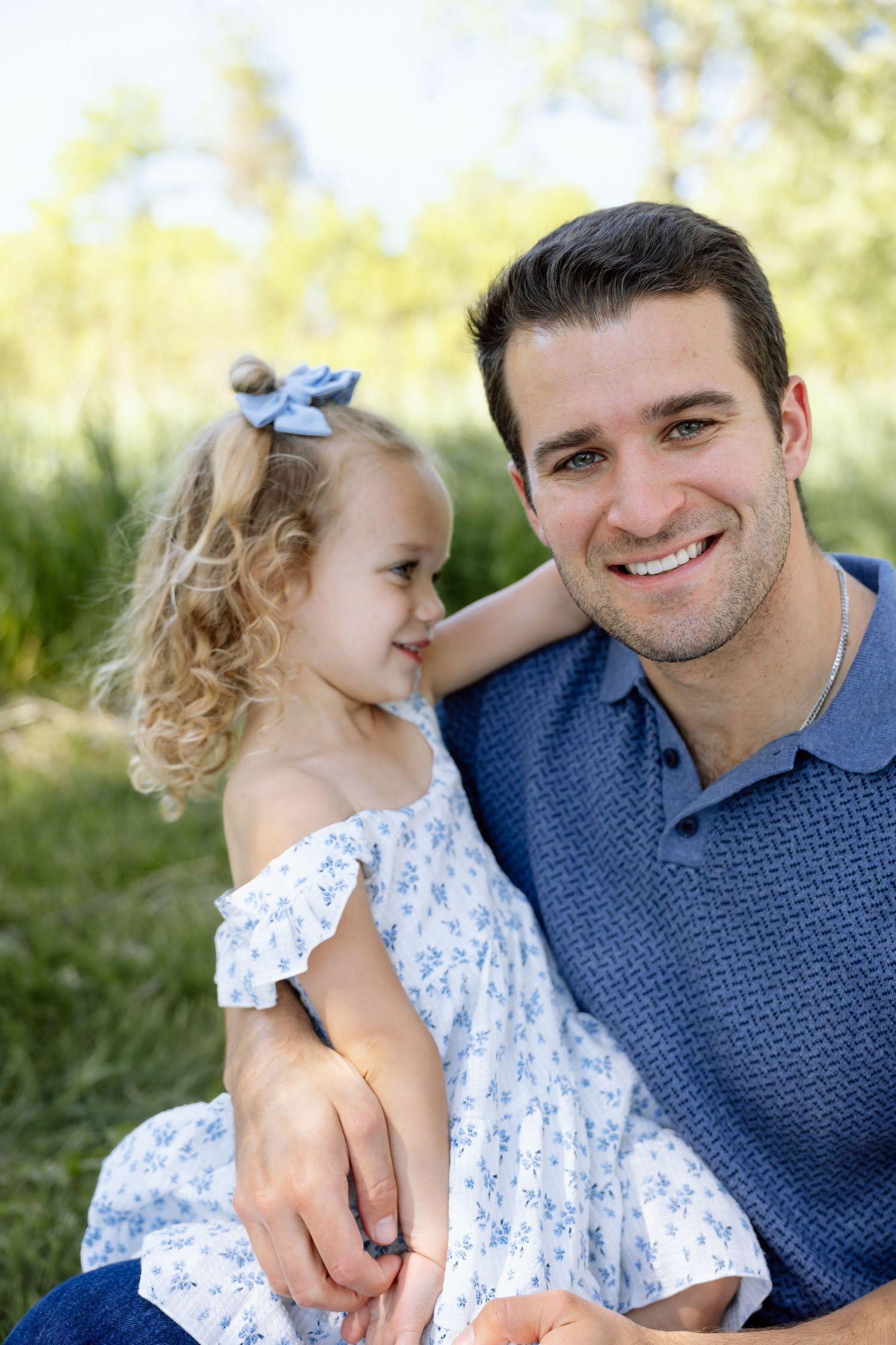Smiling father sitting outdoors with his young curly-haired daughter in a blue floral dress.