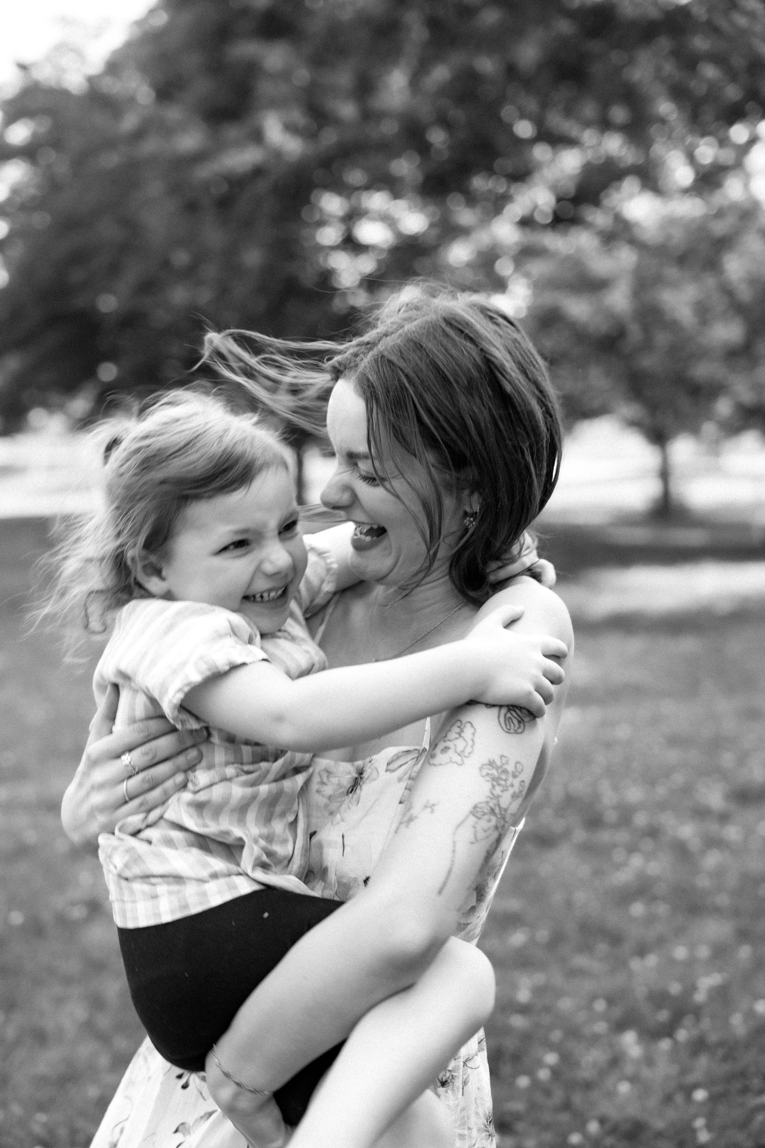 Black and white photo of a laughing mother and toddler embracing in an outdoor park.