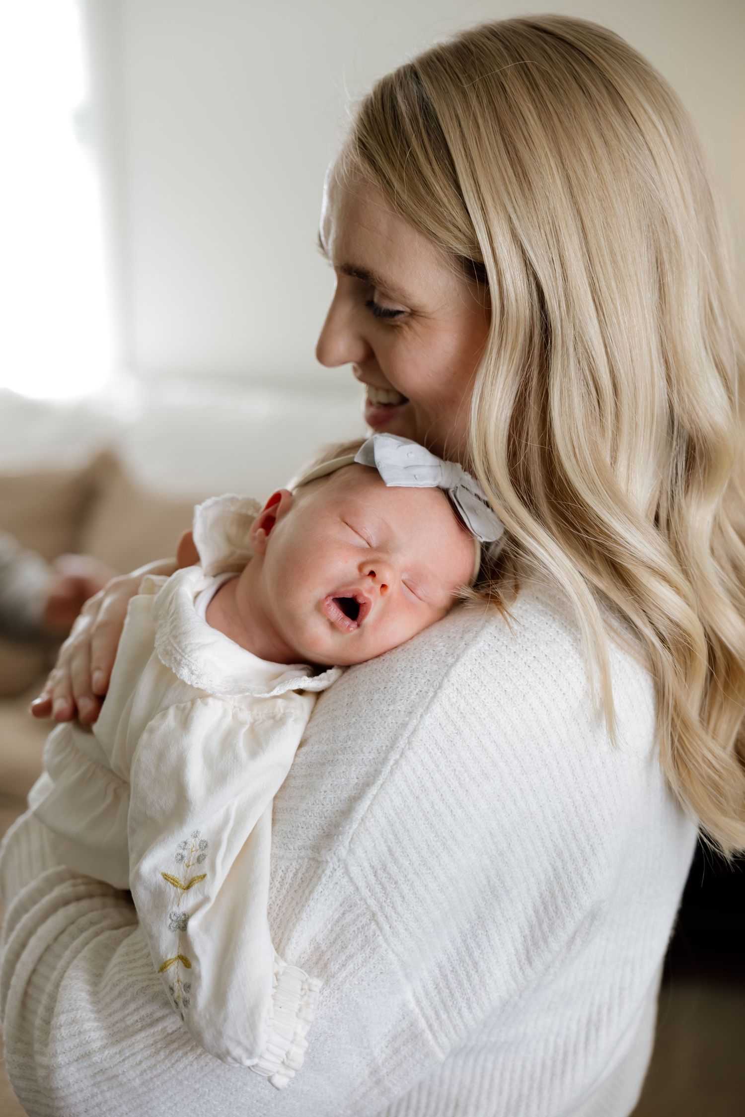 Blonde mother smiling while cradling her yawning newborn baby in white.