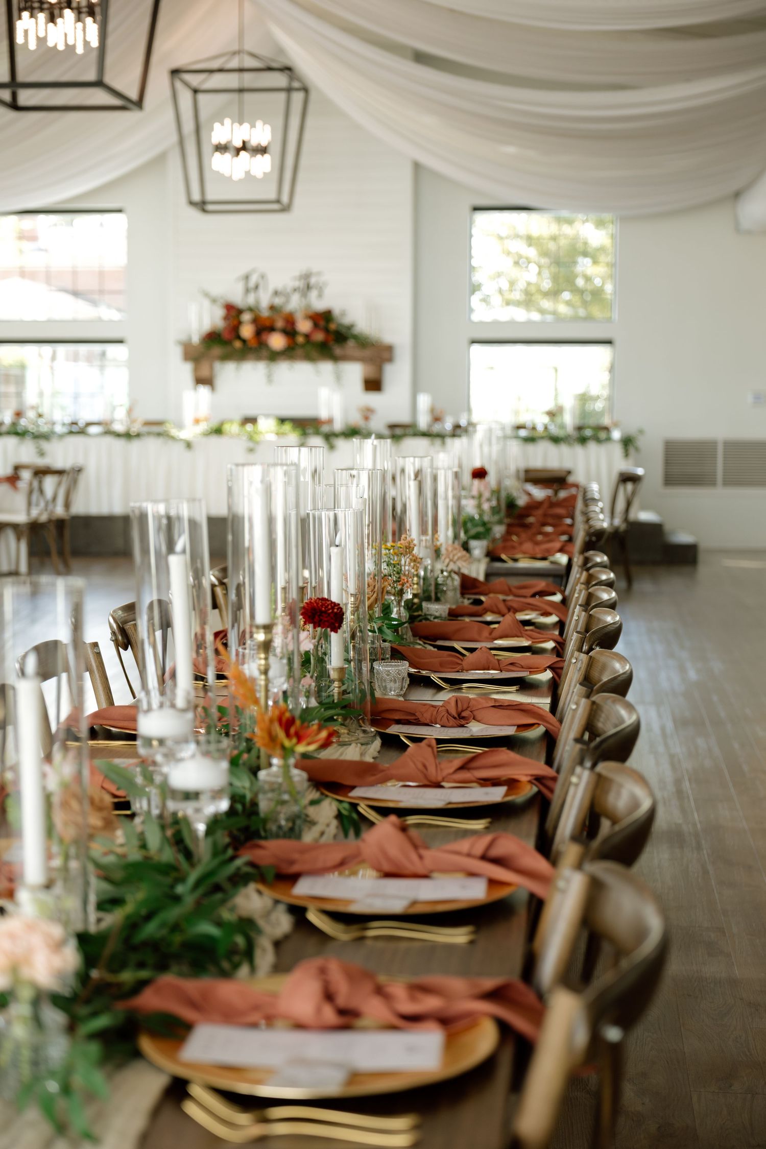 Elegant wedding reception table with terracotta napkins, gold chargers, tall glass candles, and lush floral centerpieces in a white venue.