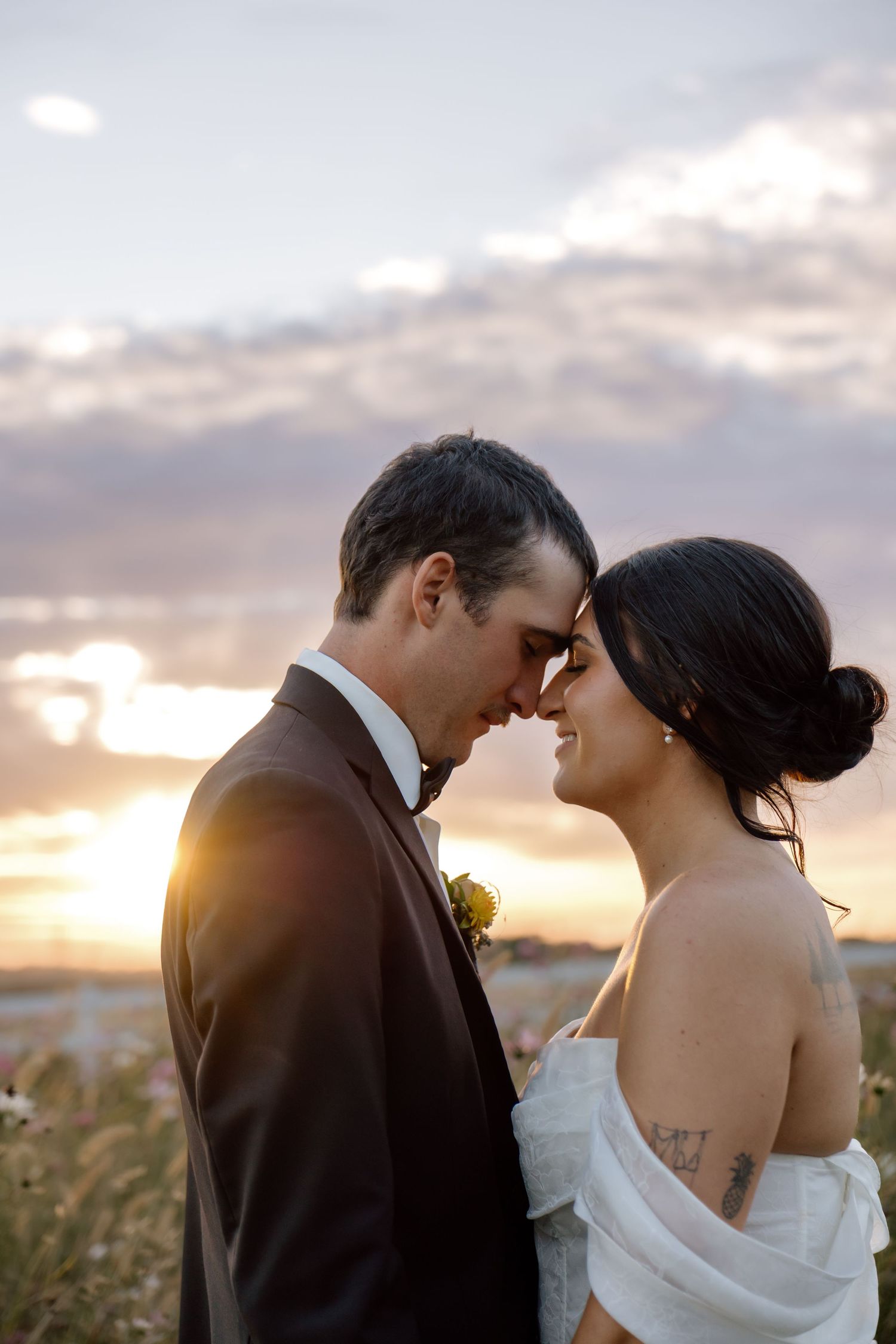 Bride and groom touch foreheads romantically in a wildflower field during a beautiful golden sunset.