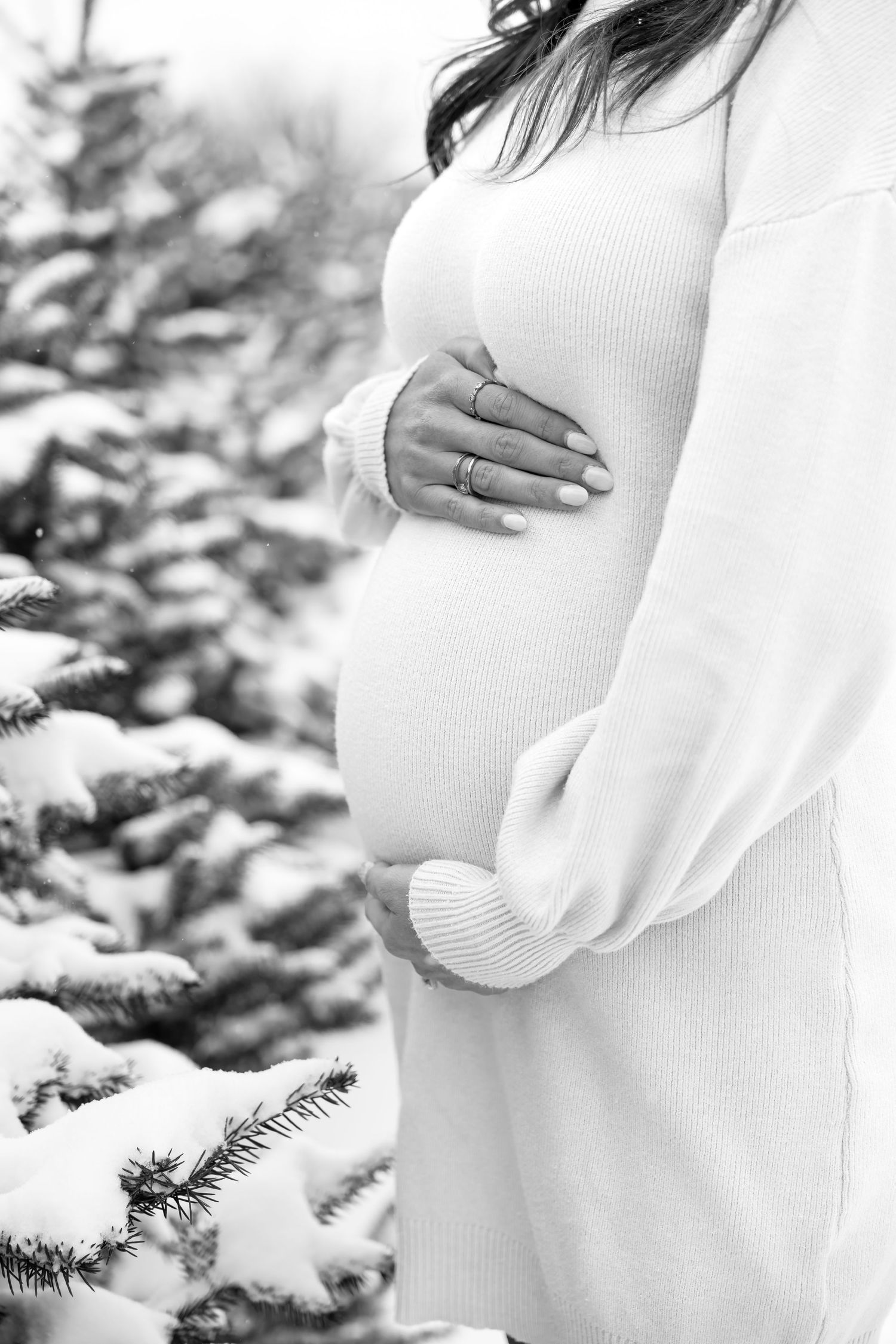 Black and white close-up of pregnant woman cradling baby bump in white knit dress outdoors.