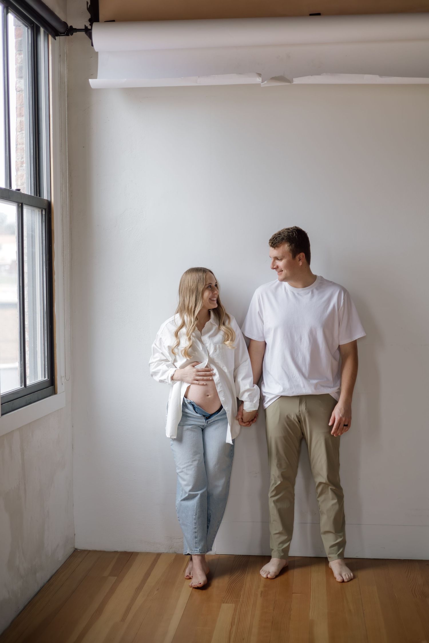 A pregnant couple holding hands and smiling at each other against a white wall in casual attire.