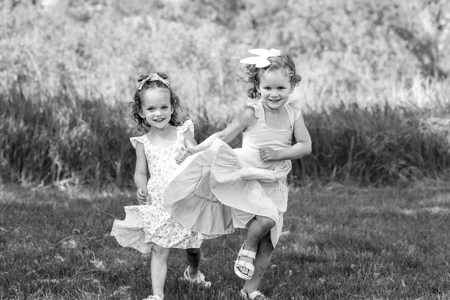 Black and white photo of two little girls in dresses running and laughing on a grassy lawn.