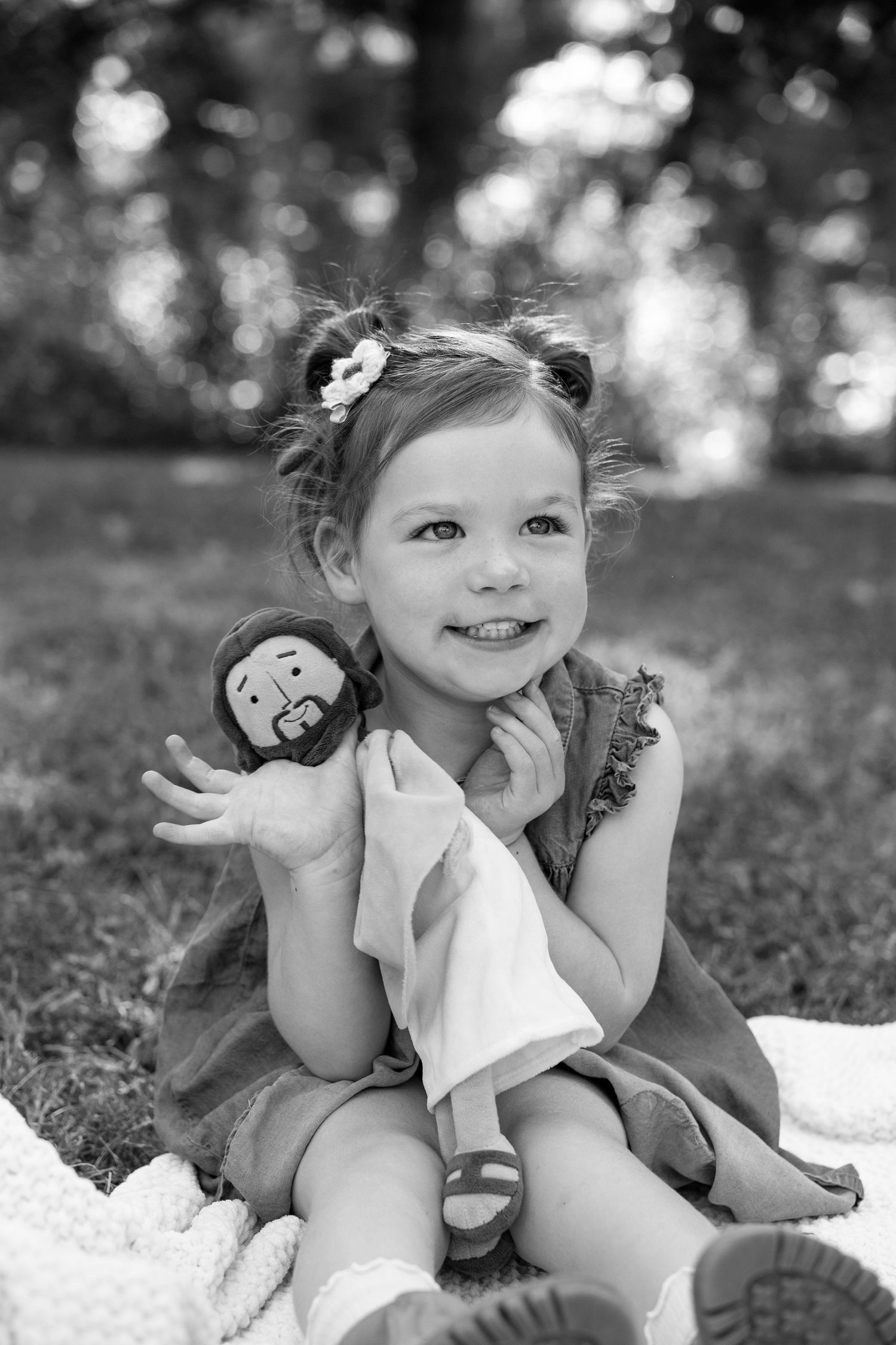 Black and white portrait of a smiling young girl holding a doll while sitting on a blanket outside.