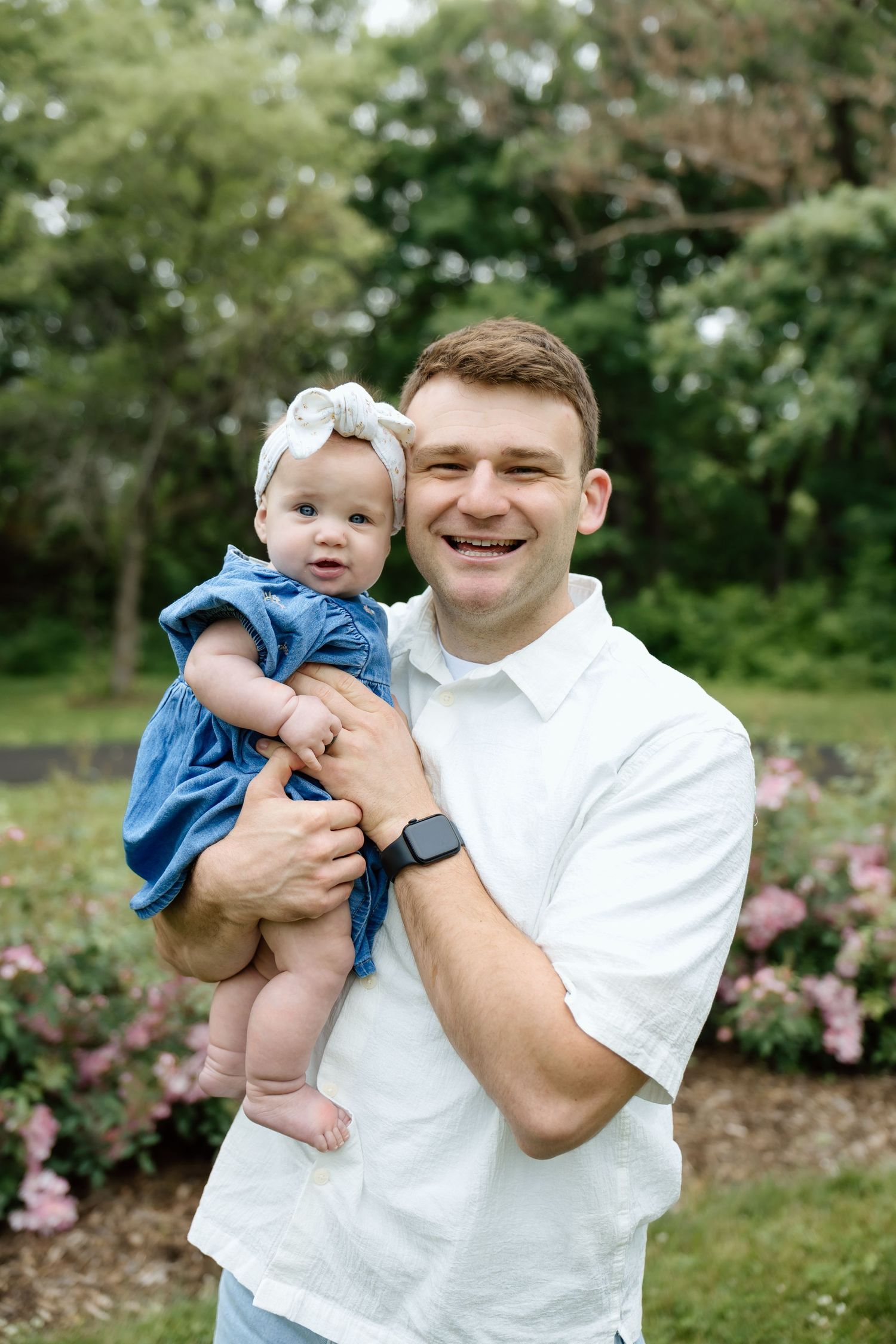 Smiling father in white shirt holding baby girl in blue dress in a flower garden.