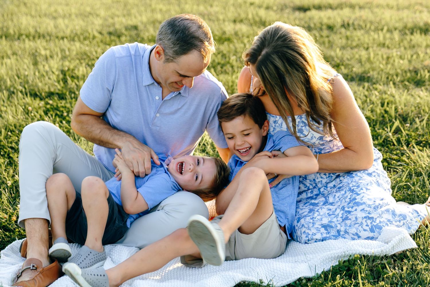 Joyful family of four laughing and playing together on a blanket in a sunny green field.