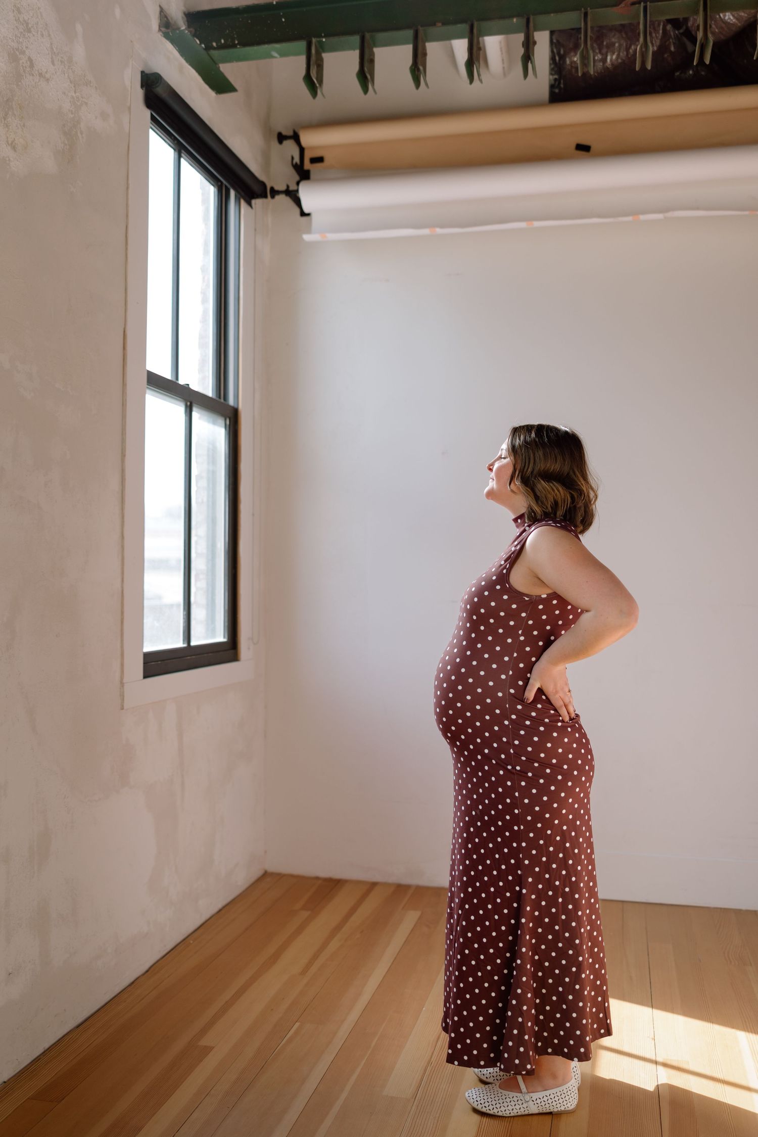 A pregnant woman in a brown polka dot dress stands by a window in a bright studio space, looking up.