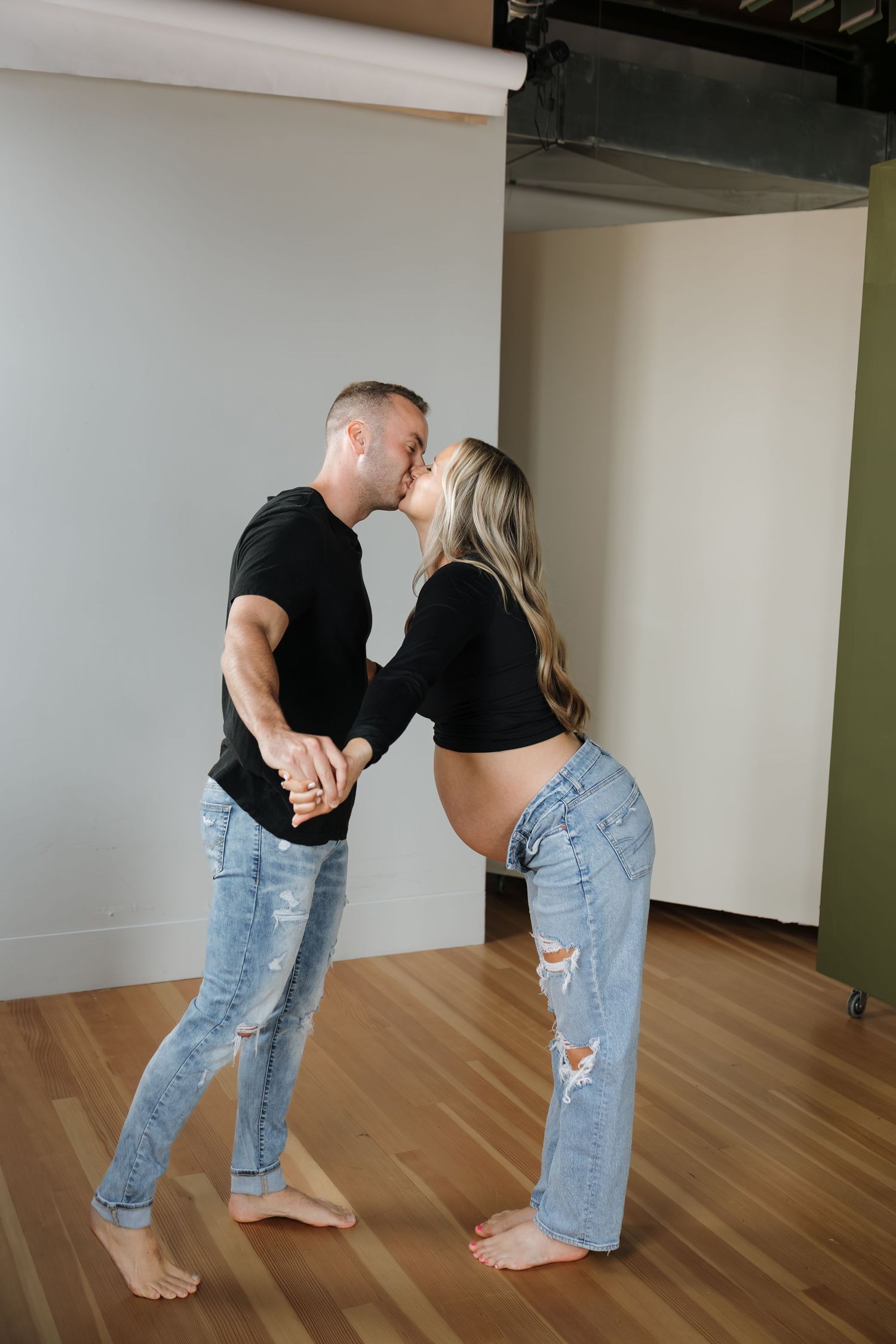 Pregnant couple sharing a kiss in a modern photography studio, woman's baby bump visible in profile.