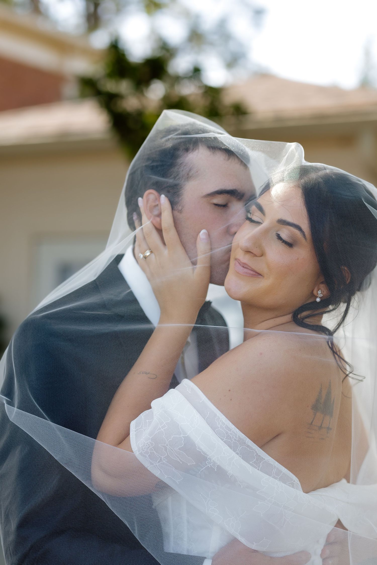 Groom kissing bride on cheek under sheer veil outdoors on wedding day.