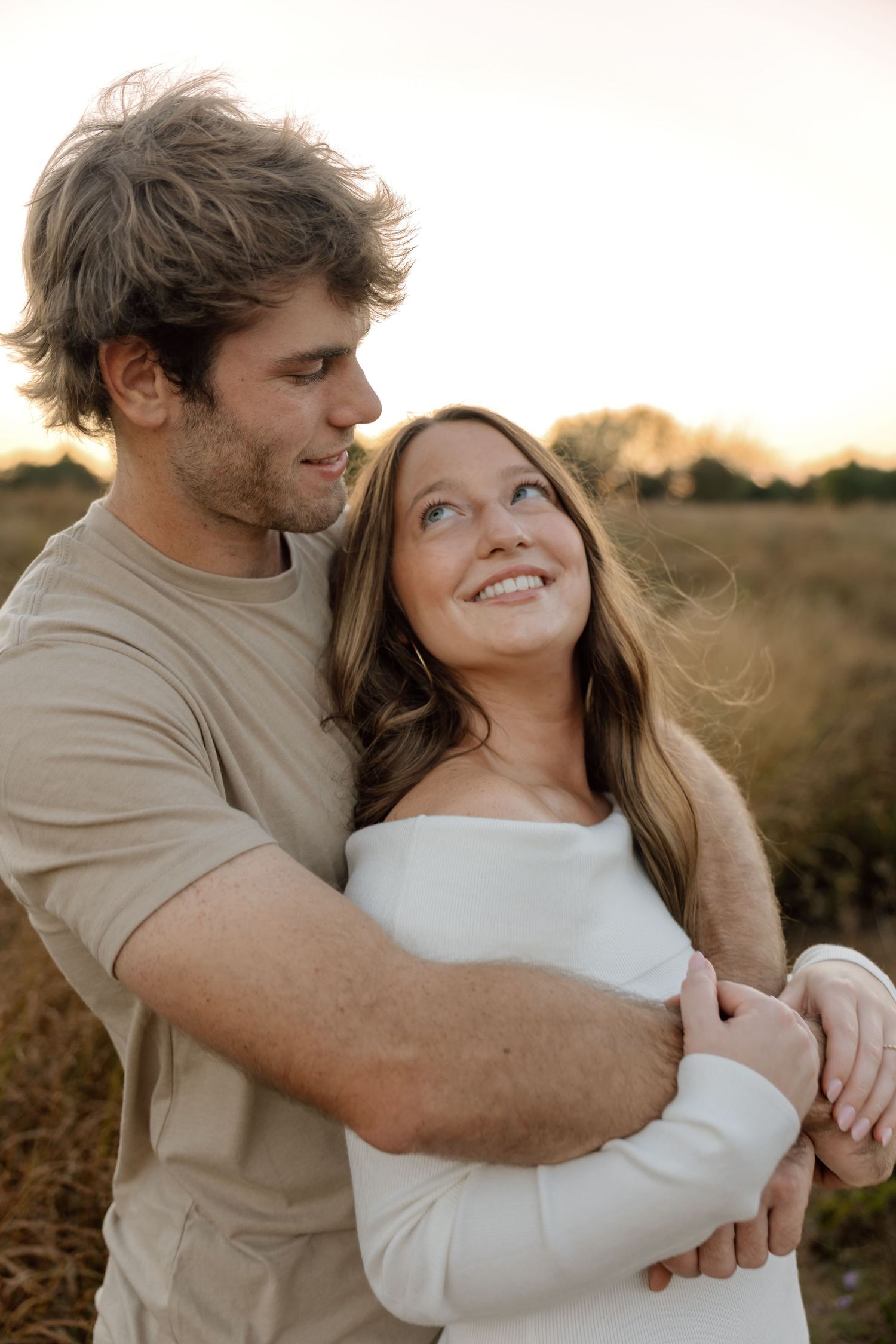Man in beige tee embracing smiling woman in white off-shoulder dress in a sun-drenched golden field.
