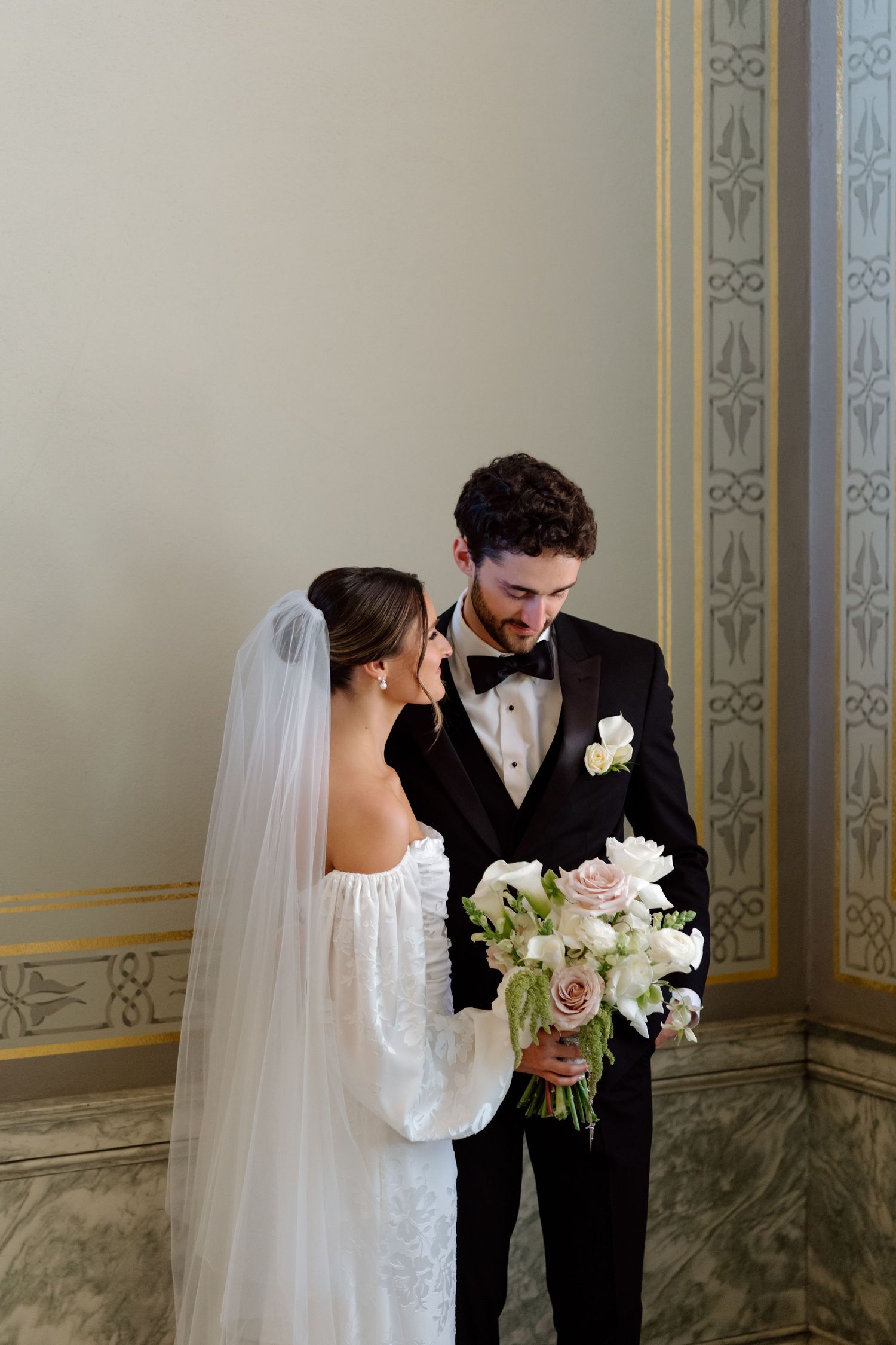 Bride in white gown and veil holds bouquet with groom in black tuxedo in elegant venue.