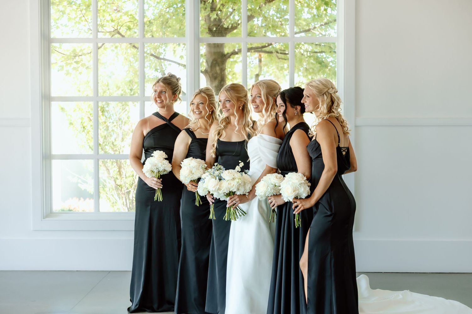 Bride in white gown poses with five bridesmaids in black dresses holding white floral bouquets indoors.
