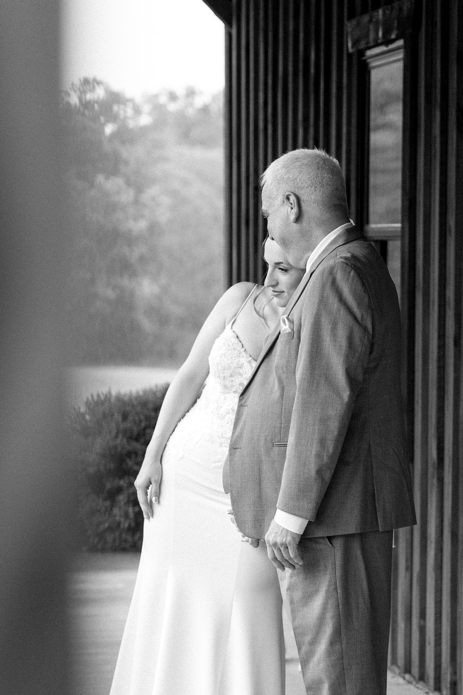 Black and white romantic photo of bride and groom holding hands leaning against column on outdoor porch.