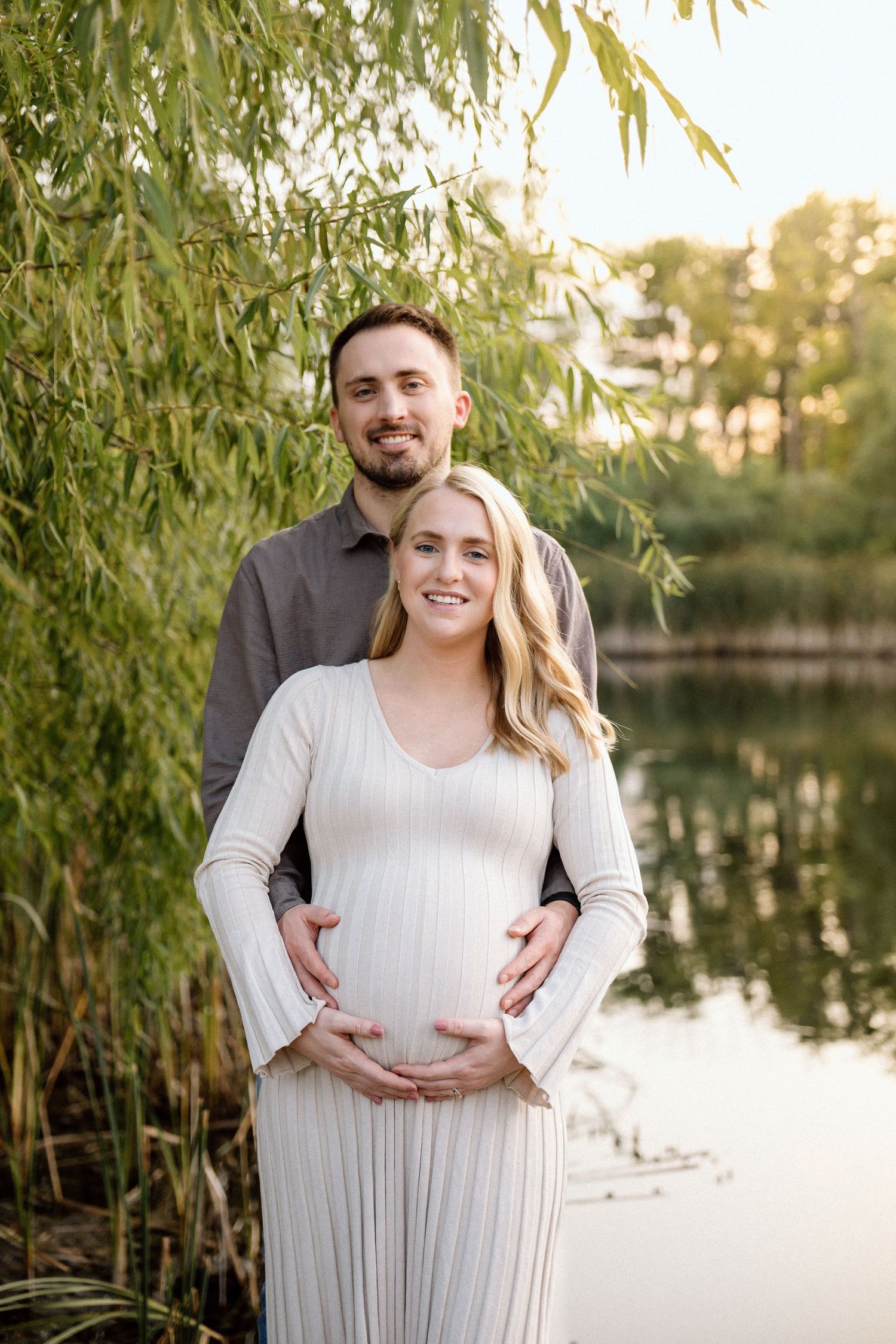 Smiling pregnant couple standing by a serene lake under willow trees, cradling baby bump in golden hour light.