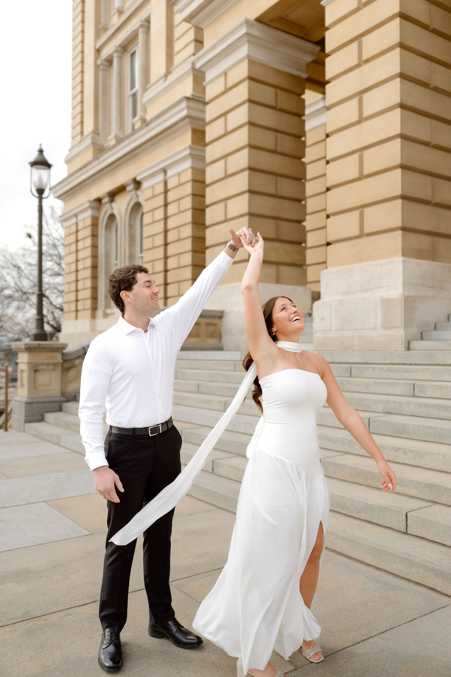 A joyful bride and groom dance outside a grand stone building, the bride in a white gown with flowing ribbon.