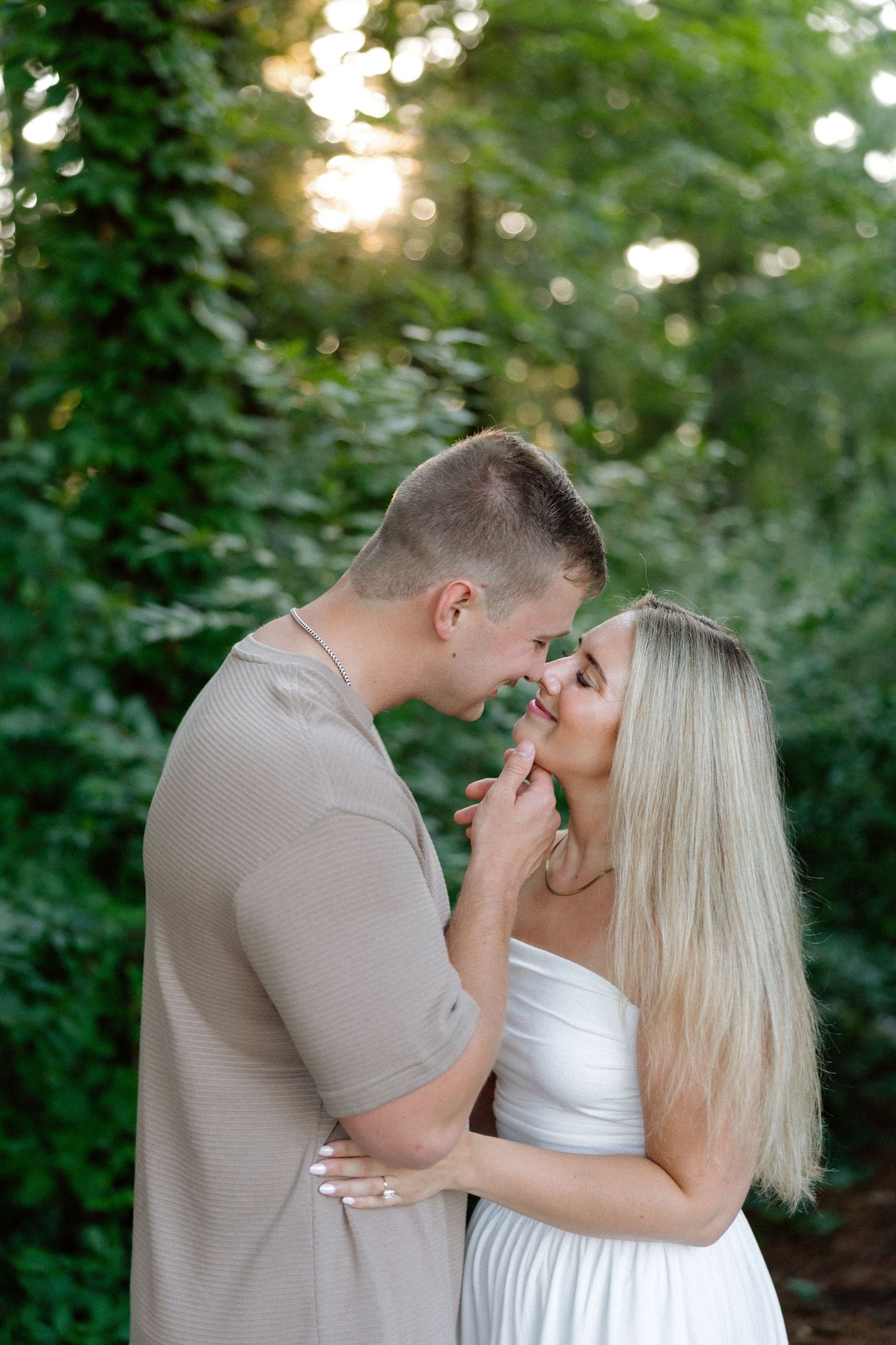 A couple sharing a romantic moment outdoors, with the man in beige and woman in white dress surrounded by lush green foliage.