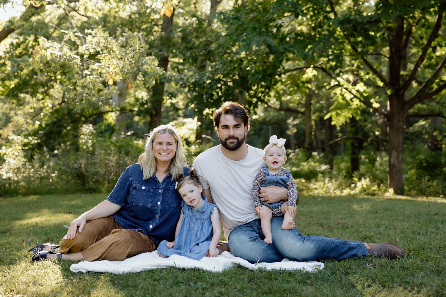 Family of four posing on white blanket in lush green park, parents with two young girls.