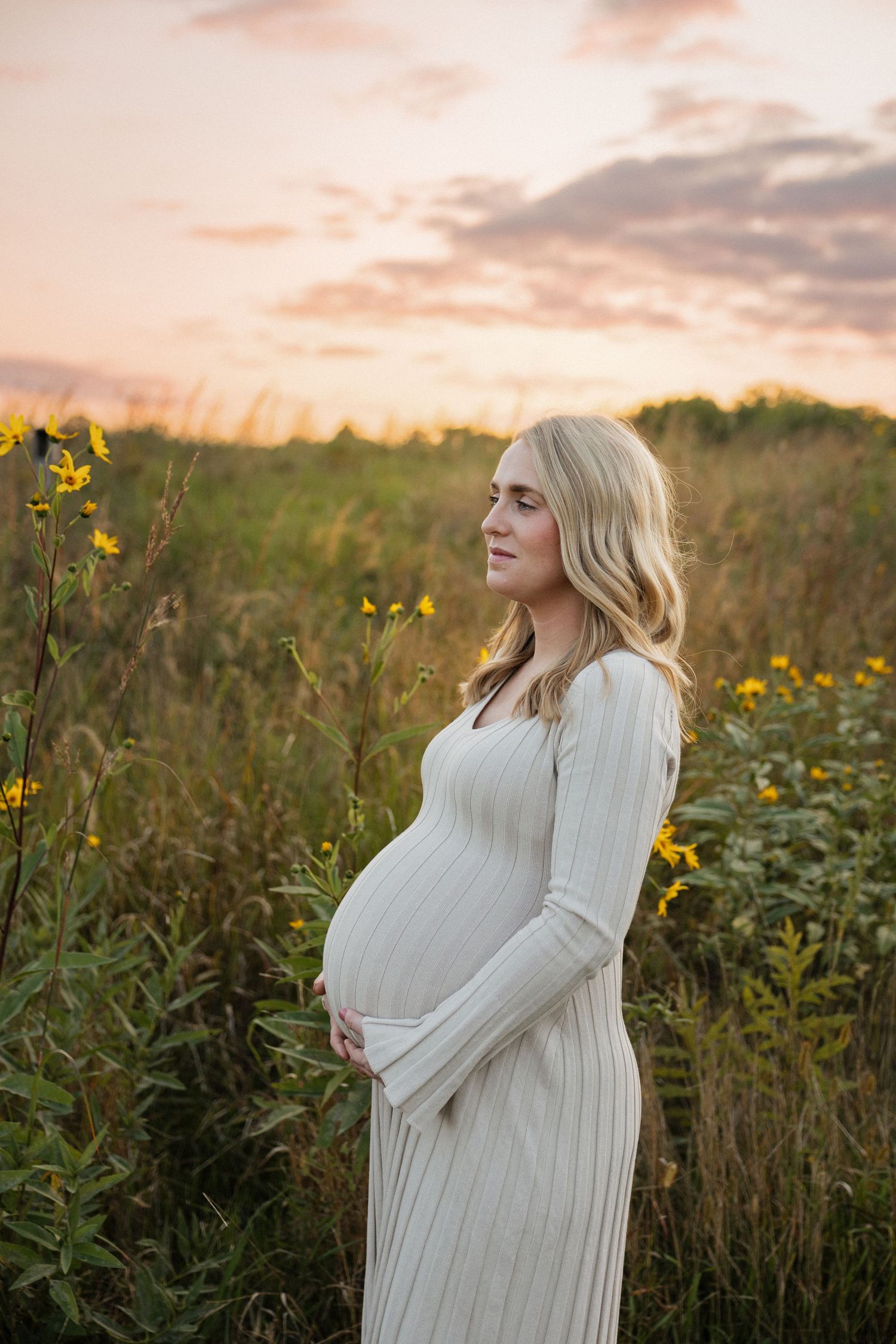 Pregnant woman in cream ribbed dress holding belly in wildflower field during vibrant pink and purple sunset.