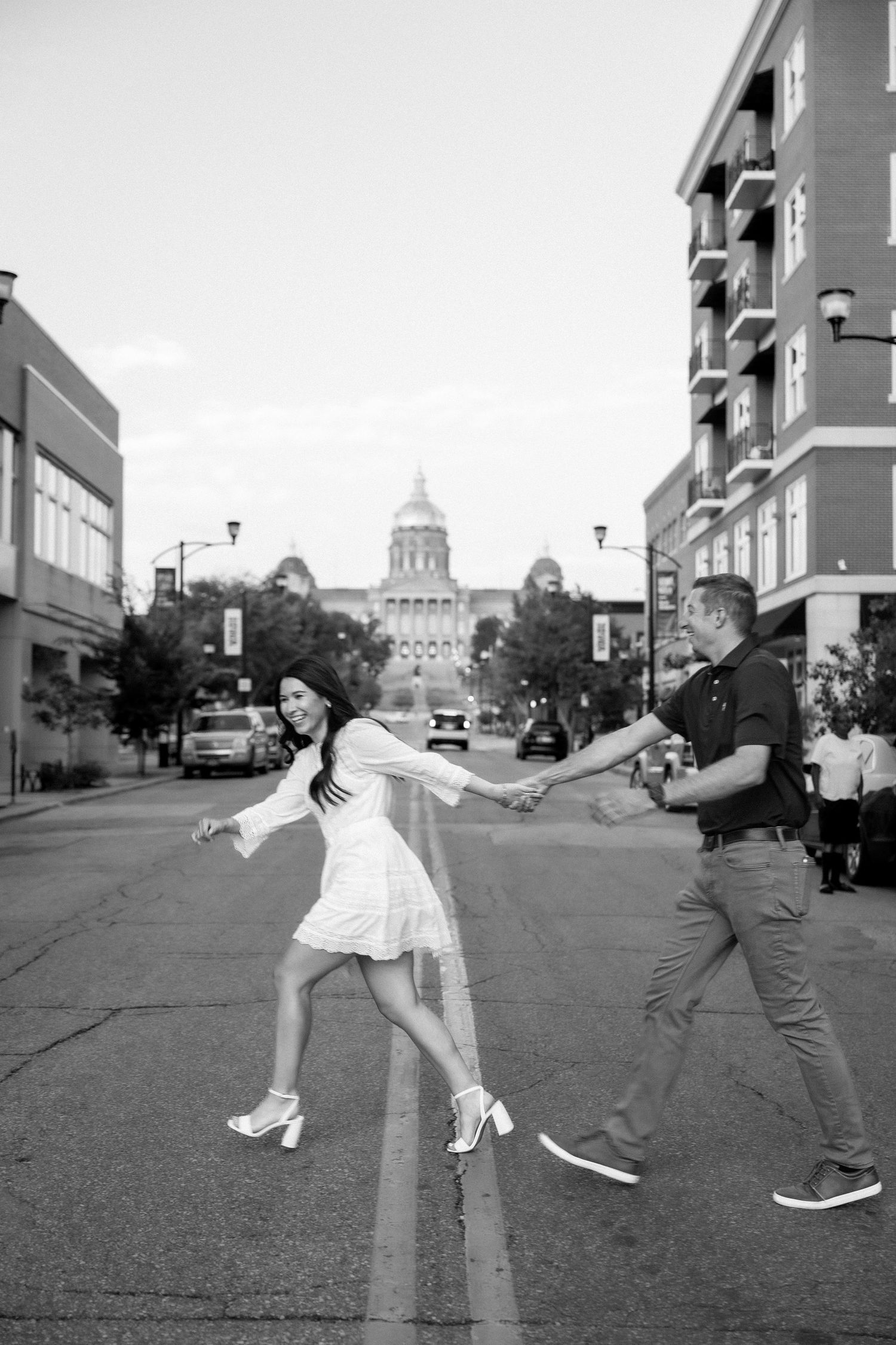 Black and white photo of a joyful couple holding hands and dancing in the street with a capitol dome behind them.