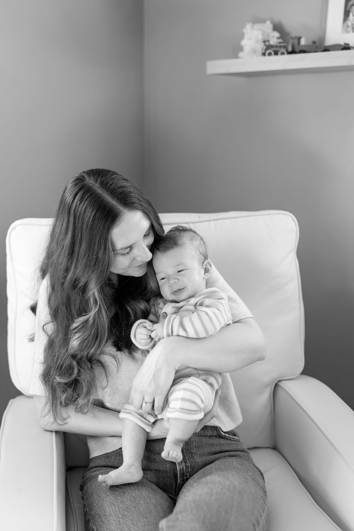 Black and white photo of a young mother sitting in a white chair, tenderly holding her smiling newborn baby.