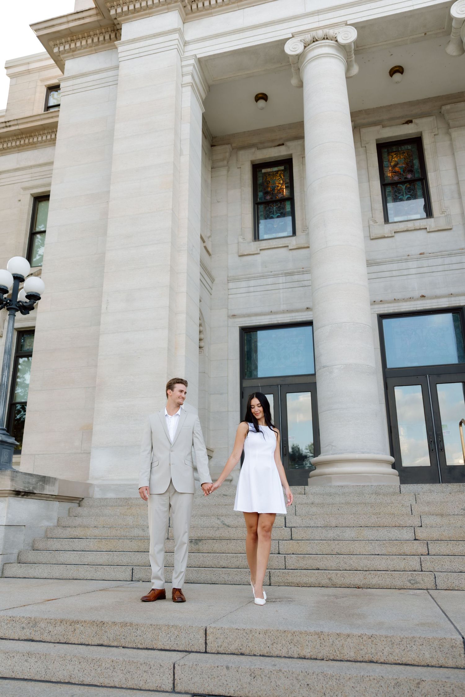 Elegant couple holding hands on steps of a grand neoclassical building, man in gray suit, woman in white dress.