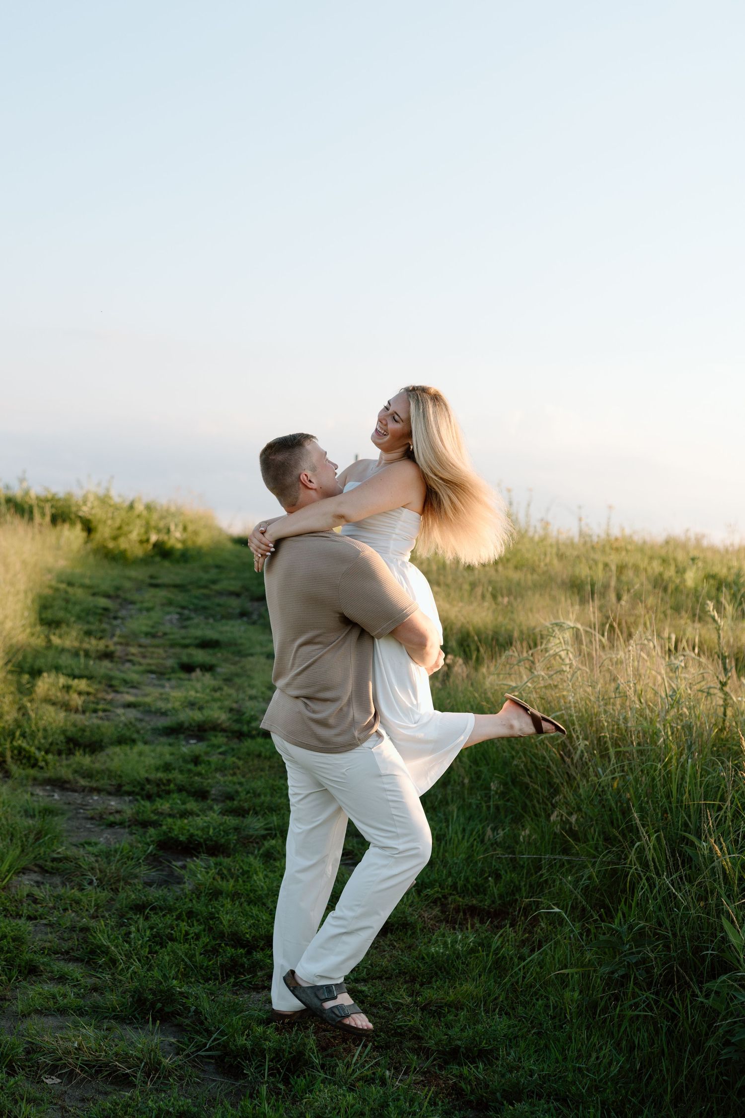 A man lifts a smiling blonde woman in a white dress outdoors on a green field path at sunset.