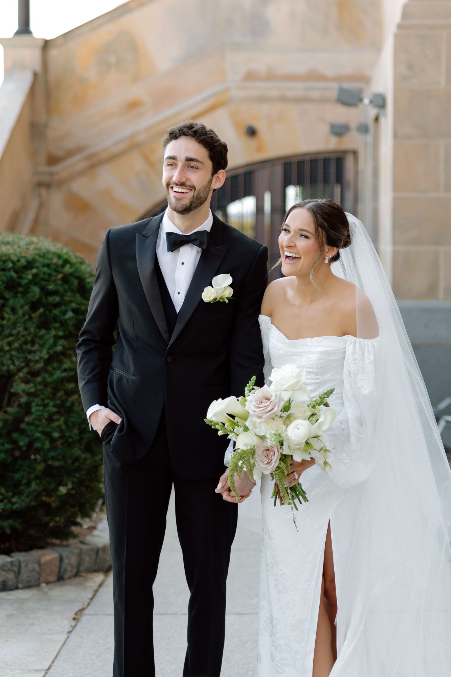 Happy bride and groom laugh together outdoors, she holds white floral bouquet, he wears classic black tuxedo.