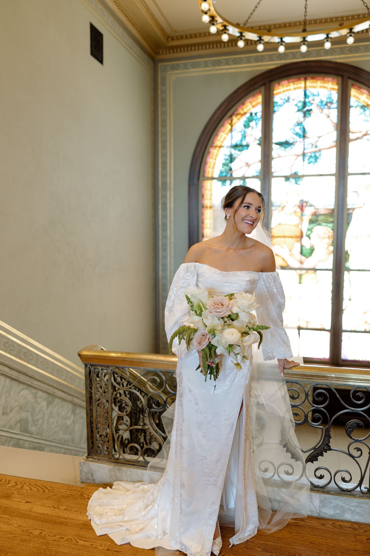 Bride in off-shoulder lace wedding gown holding white bouquet stands by stained glass window on balcony.