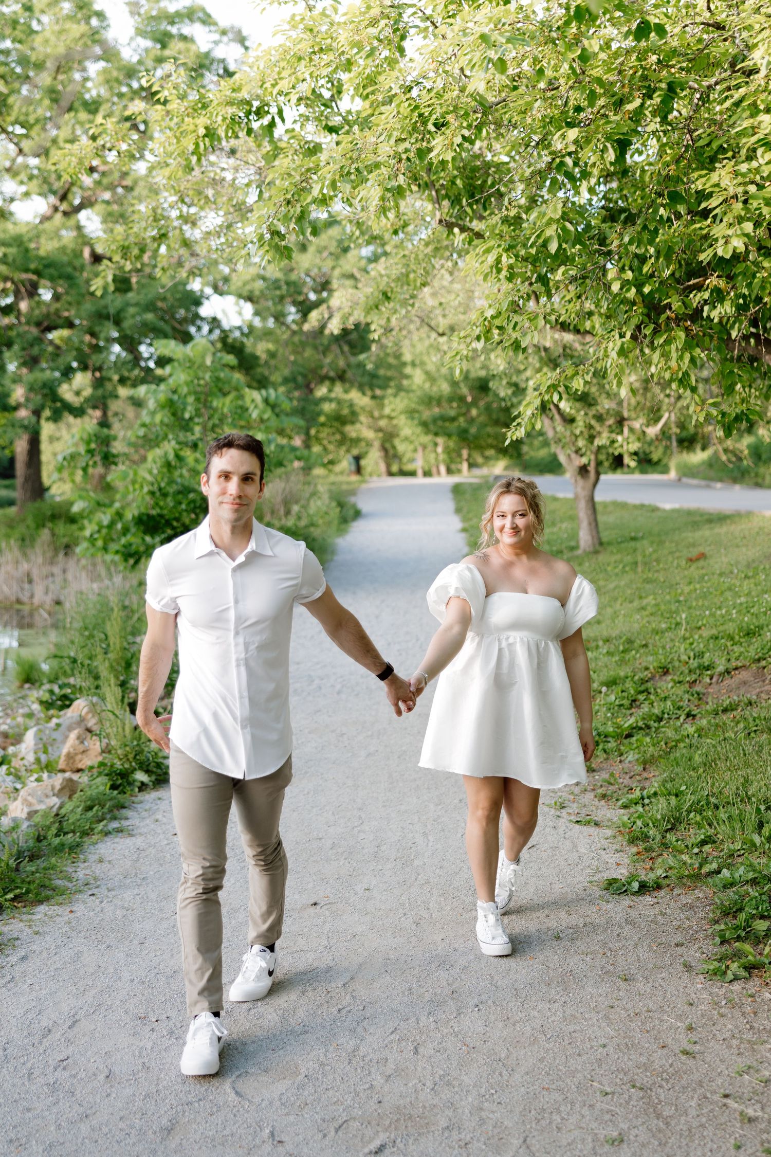 Couple in matching white outfits holding hands while walking on a scenic park path surrounded by lush green trees.