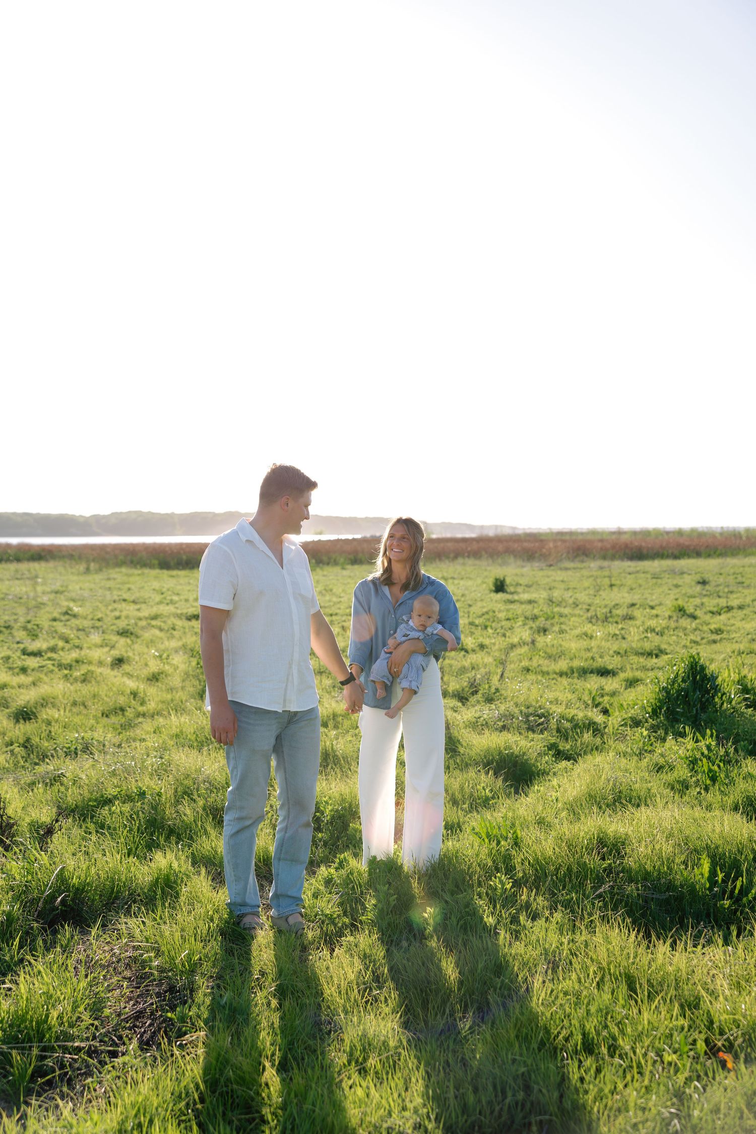 A young couple holding hands in a sunny green field, with the woman cradling a newborn baby outdoors.