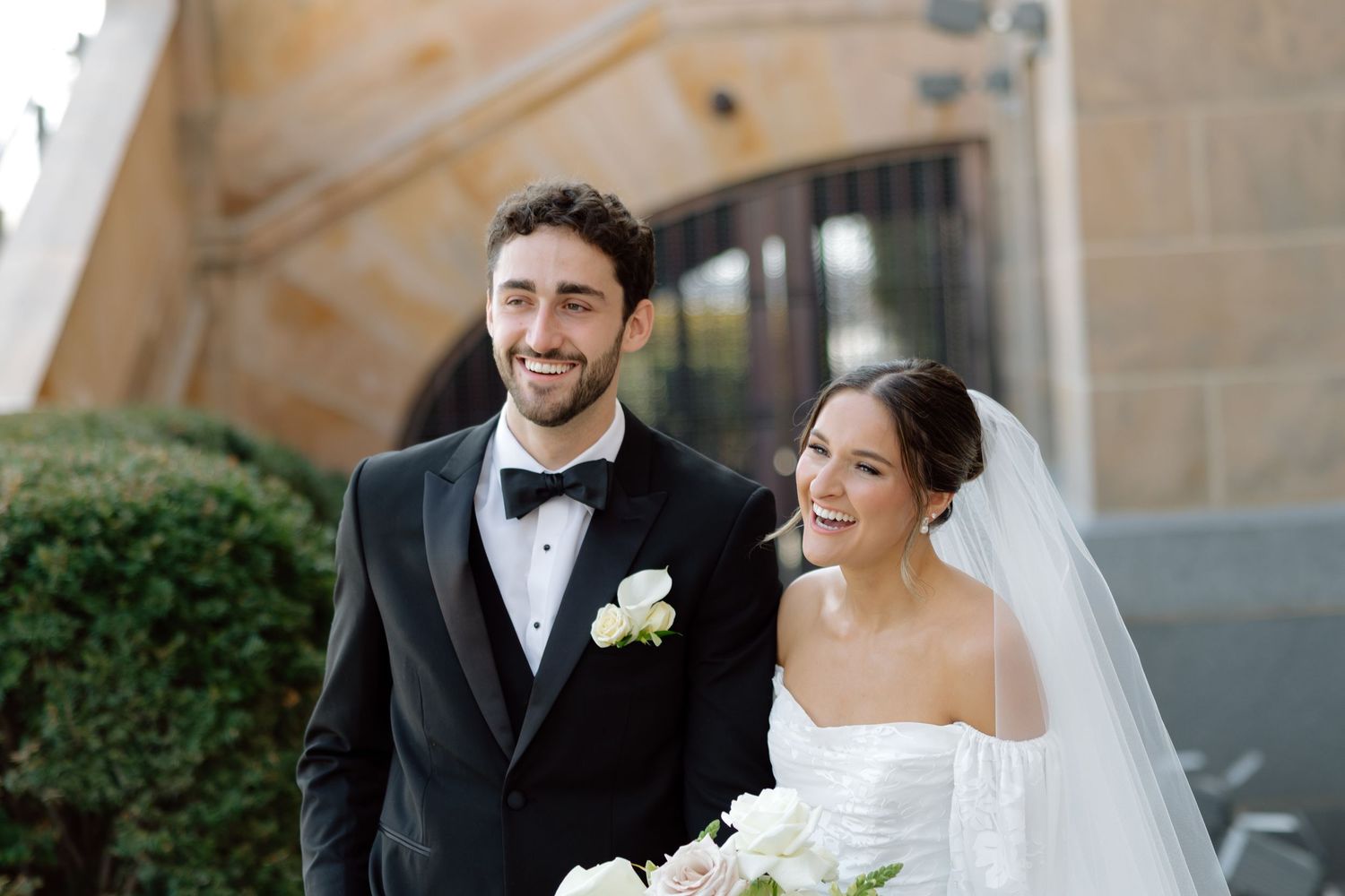 Happy bride and groom smiling outside church on wedding day, groom in tuxedo, bride in white gown with veil.