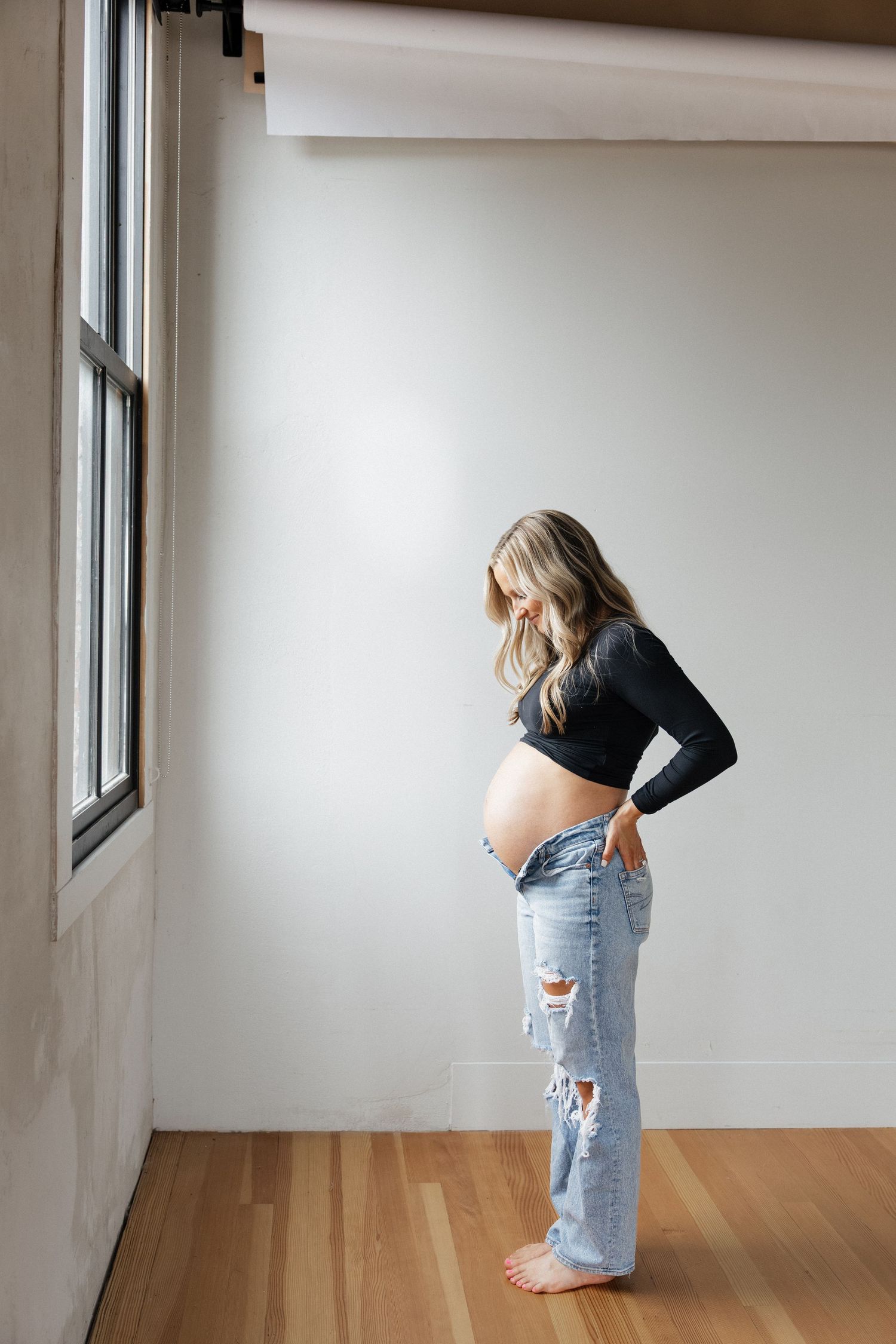 Pregnant woman in black crop top and ripped jeans standing by window, looking down at her bare baby bump.