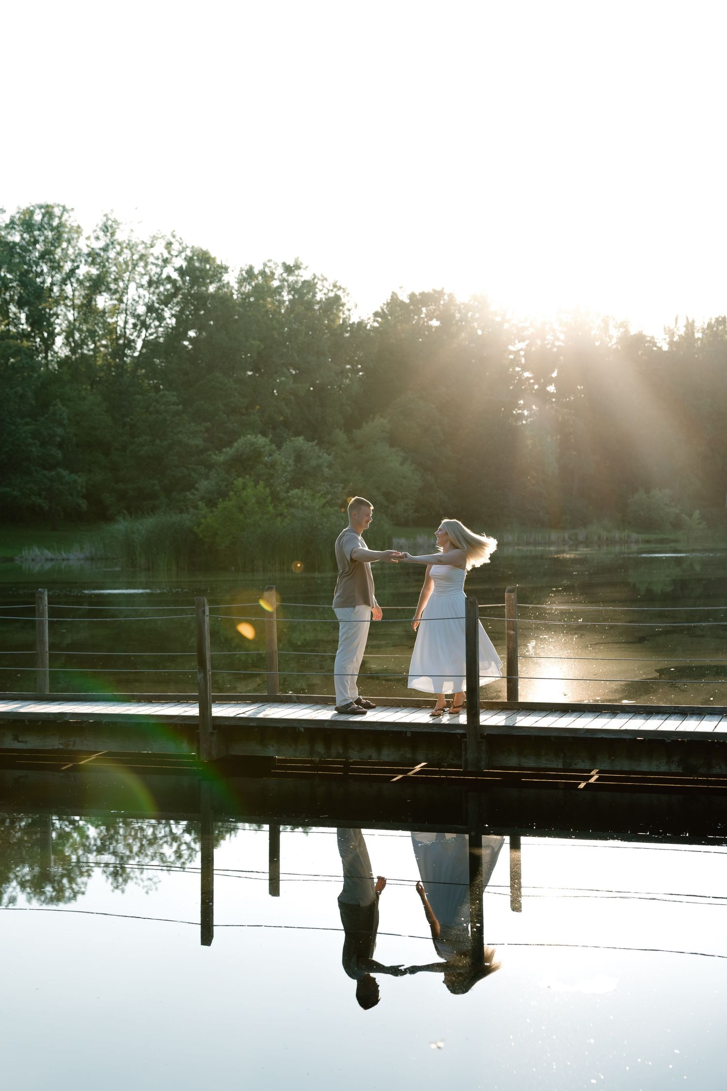 Couple holding hands on a wooden dock at sunset, with their reflection visible in the calm lake water surrounded by trees.