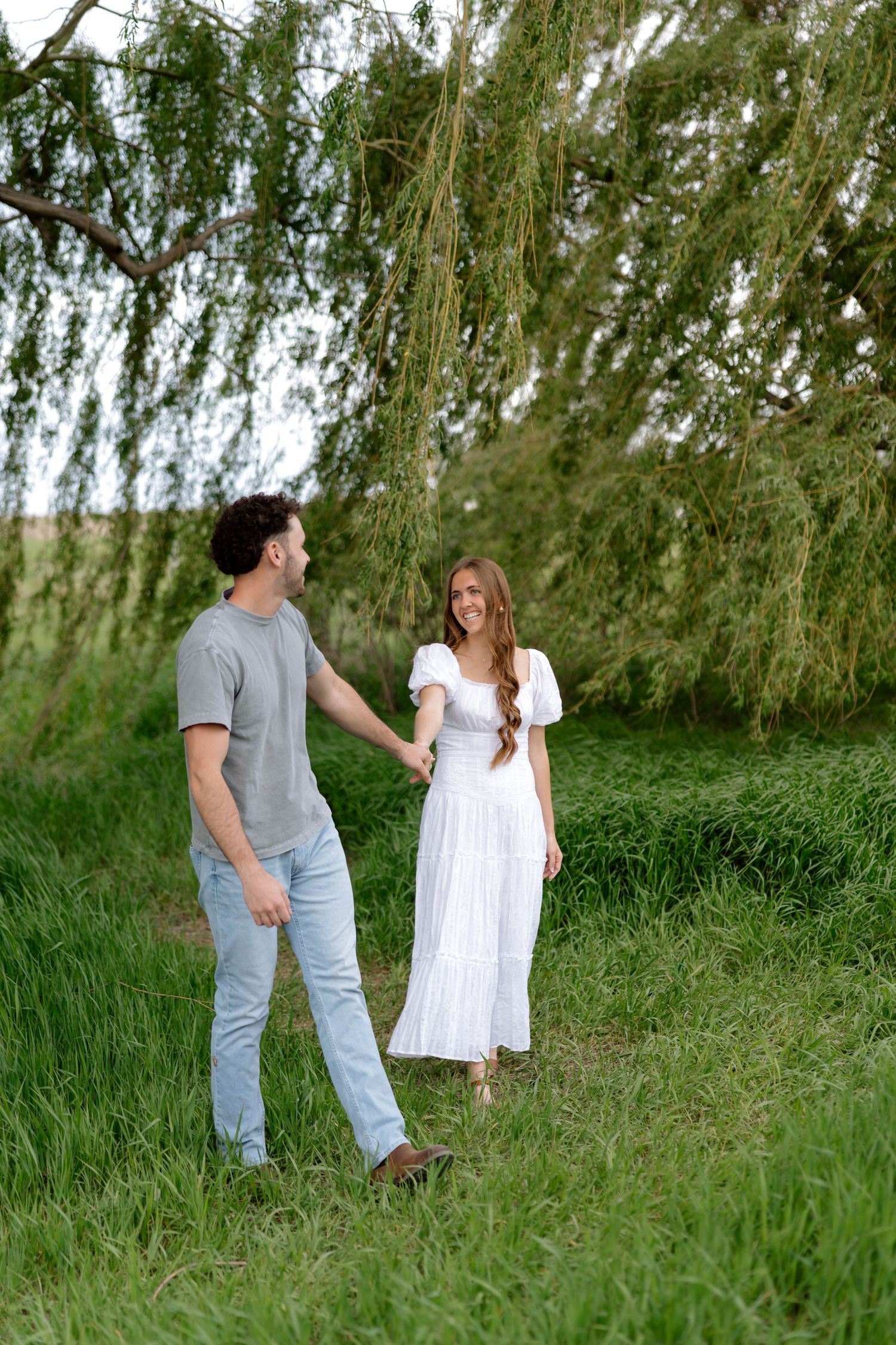 A couple holds hands in a lush green field under a weeping willow, woman in white dress, man in gray shirt.