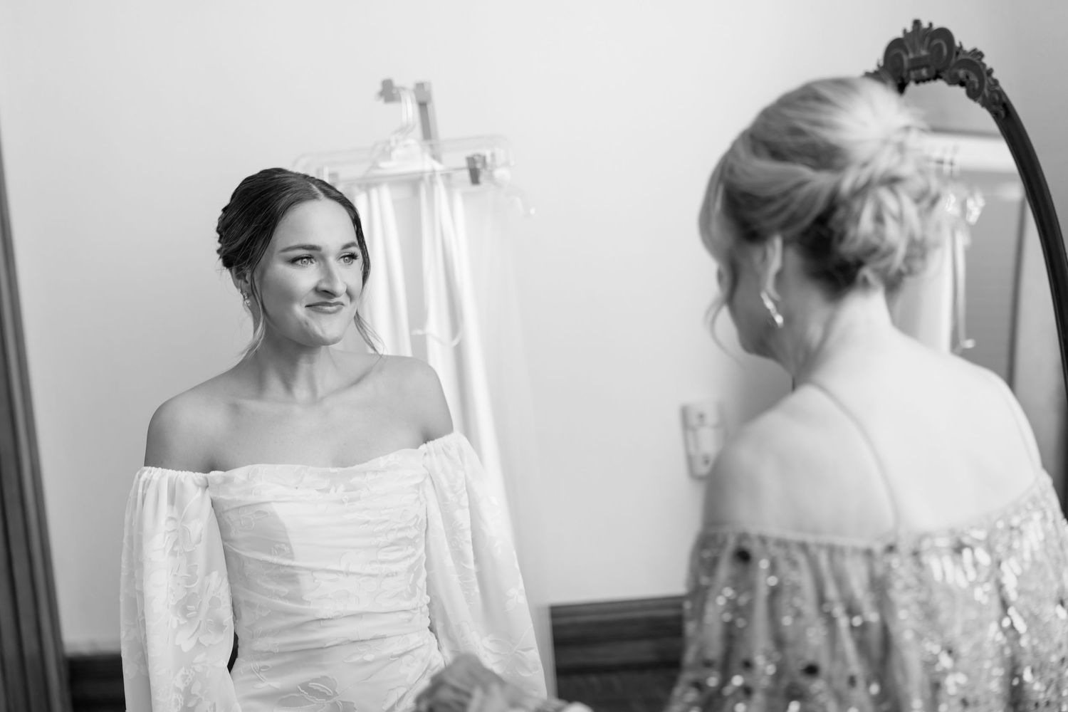 Bride in off-shoulder white wedding gown smiling at reflection in mirror in black and white photo.