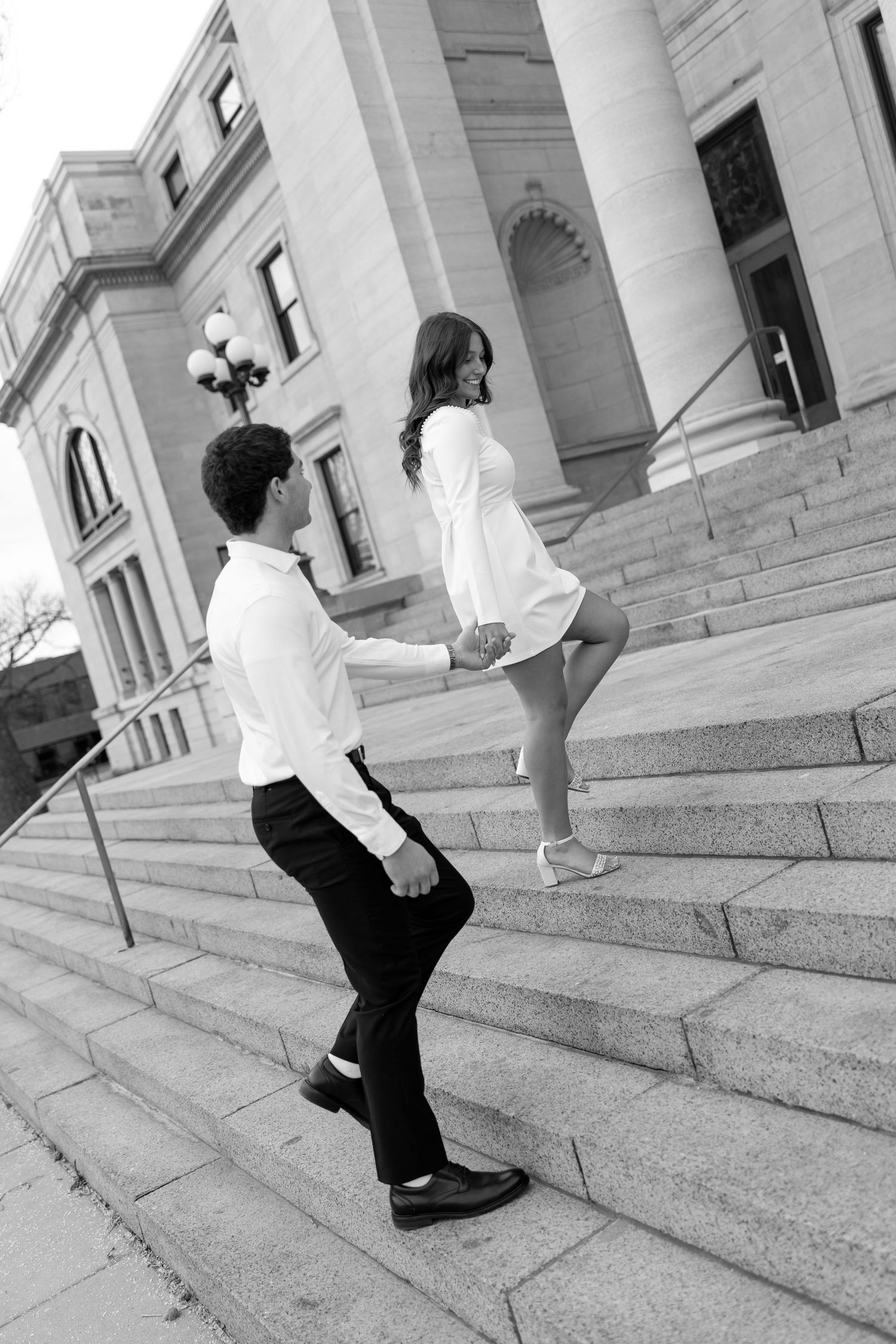 Black and white photo of couple holding hands while climbing grand stone steps of a classical building together.