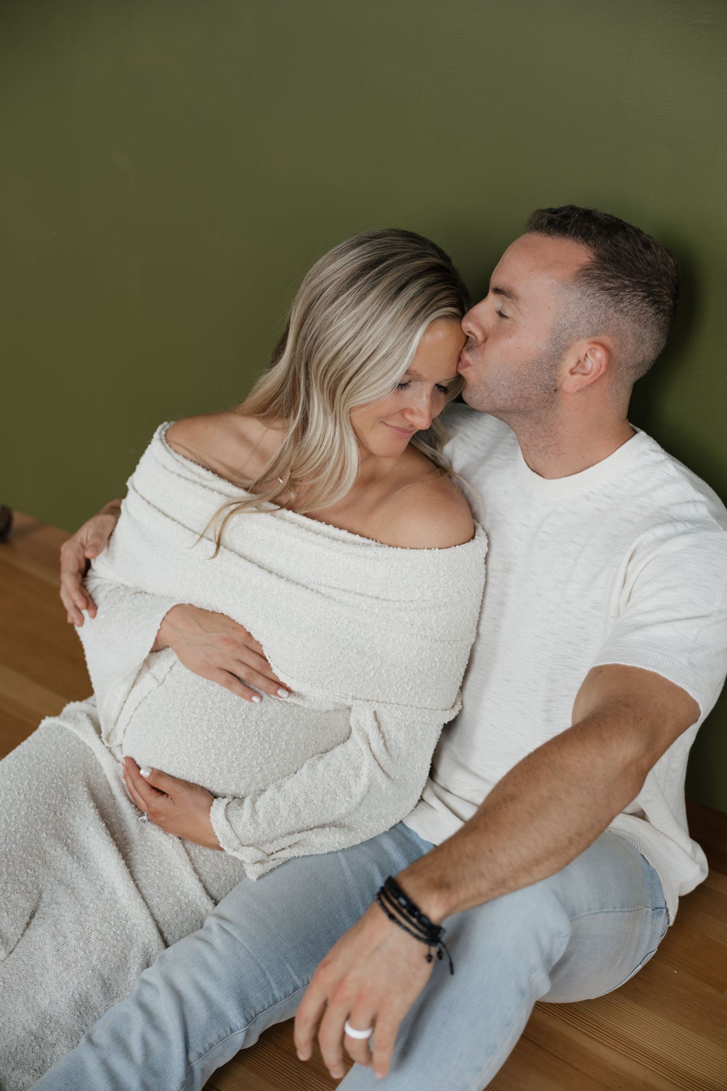 Man kissing pregnant womans forehead while sitting on floor against olive green wall.