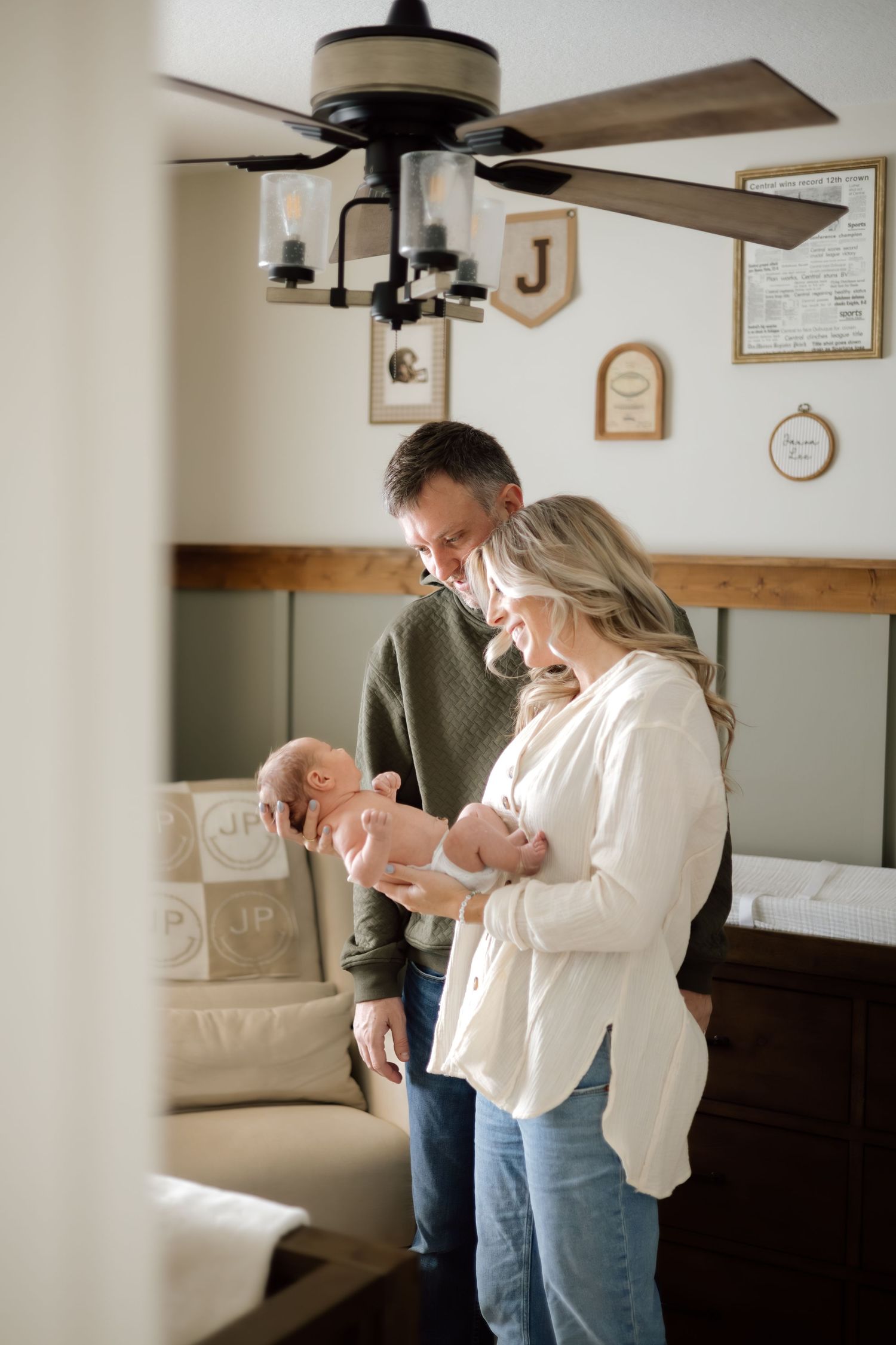 New parents in neutral tones smile down at their newborn baby in a cozy decorated nursery.