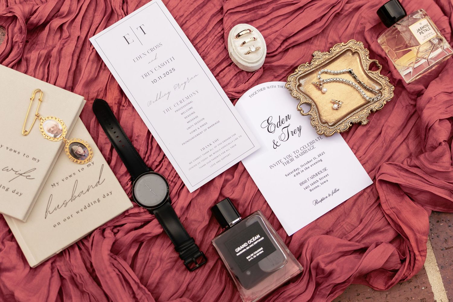 Flat lay of wedding stationery, perfume bottles, watch, and accessories on a rich burgundy fabric backdrop.