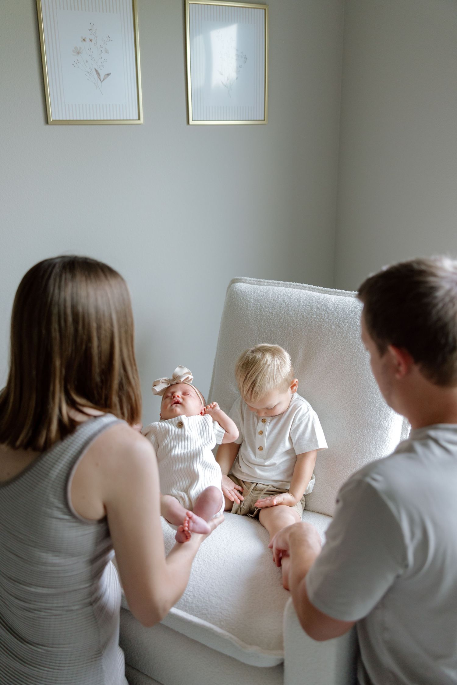 Parents watching toddler interact with newborn baby sister on white glider chair in nursery.