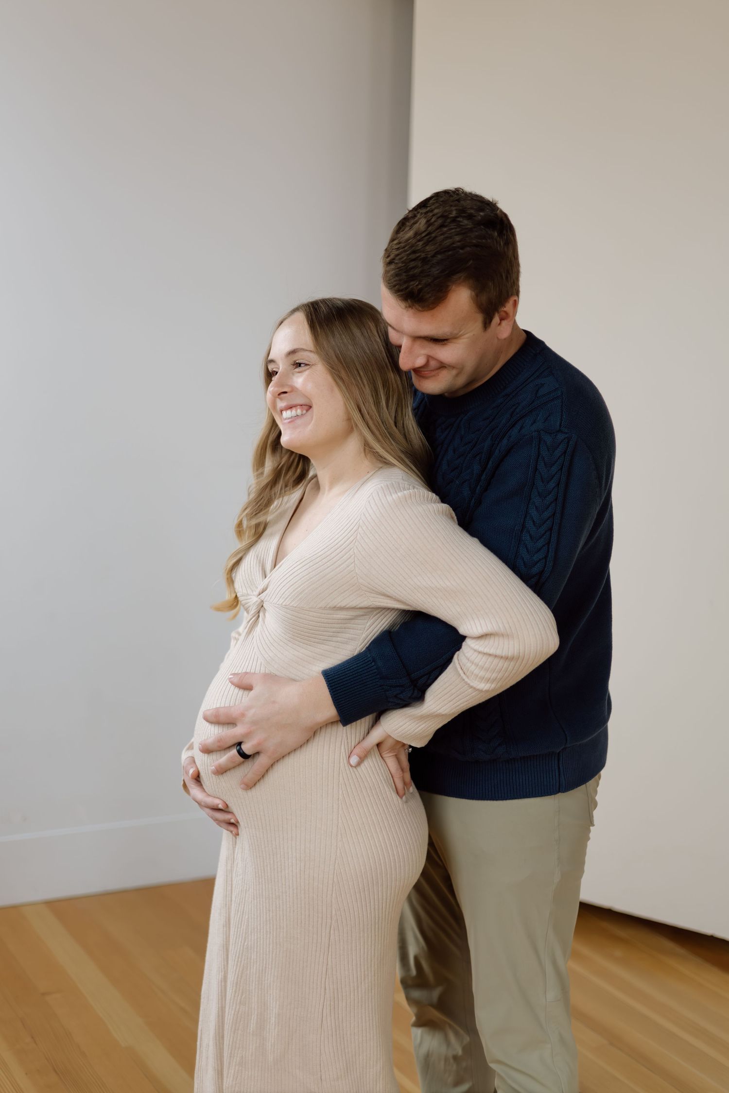 A smiling pregnant woman in a beige dress embraced from behind by her partner in a navy sweater.