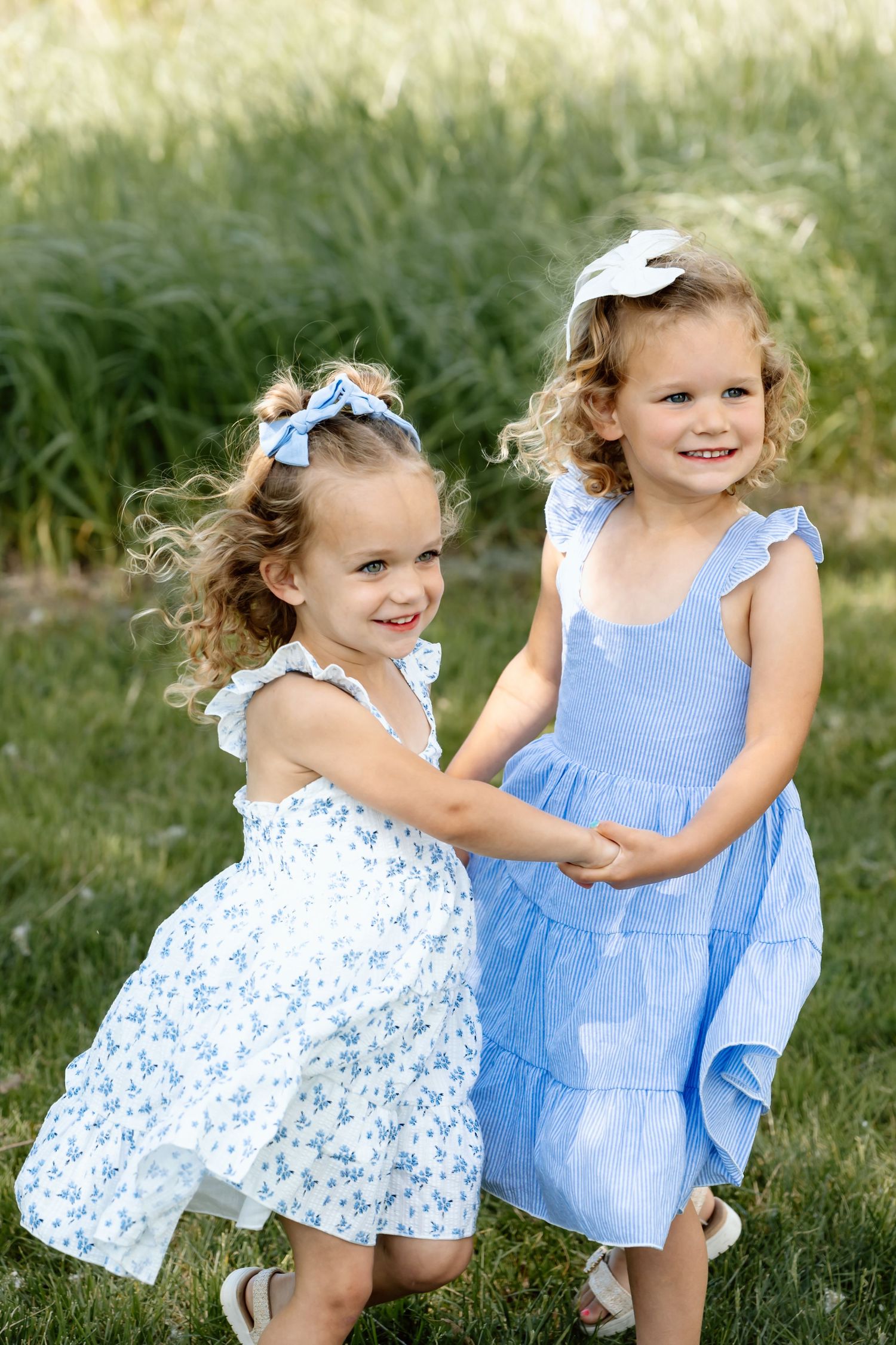 Two young girls in blue and white dresses holding hands and smiling outdoors.