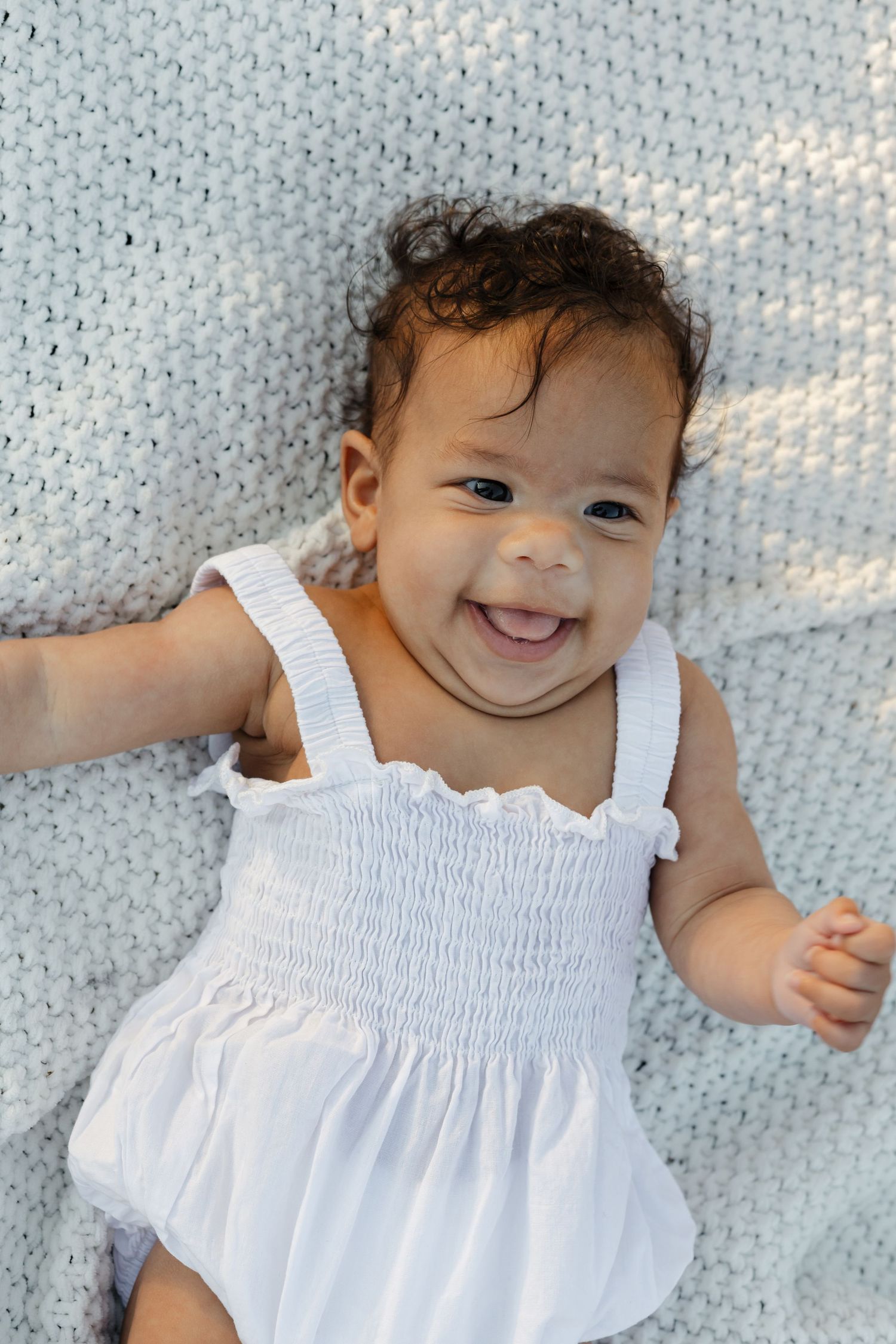 Smiling baby girl in white dress lying on a gray knit blanket.