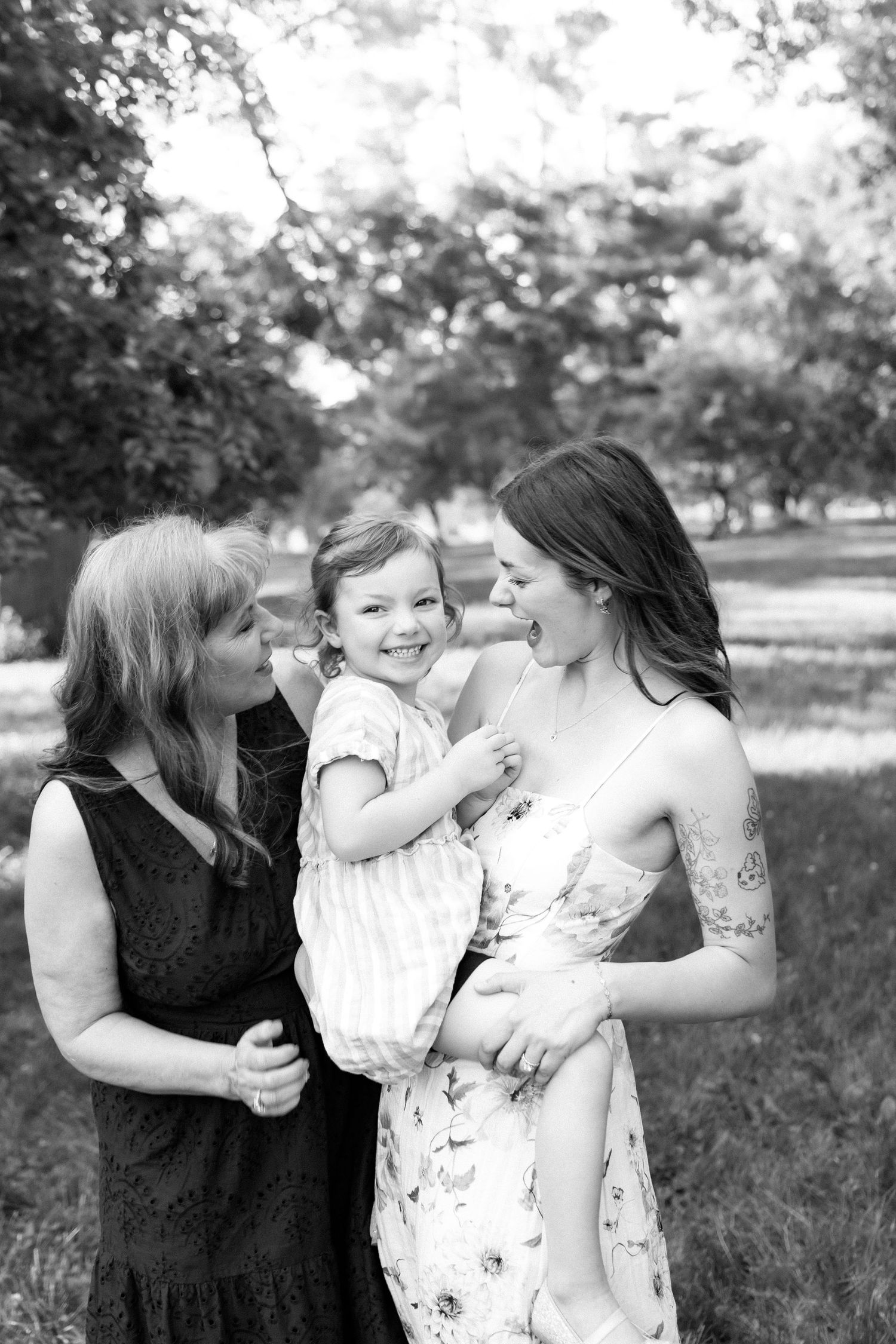 Black and white photo of a grandmother and mother smiling at a laughing toddler girl outdoors.