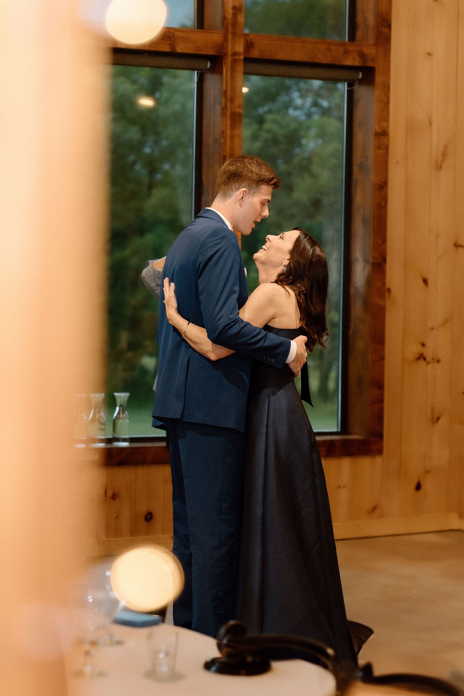 A couple shares an intimate first dance in a warmly lit wooden wedding venue with large windows.
