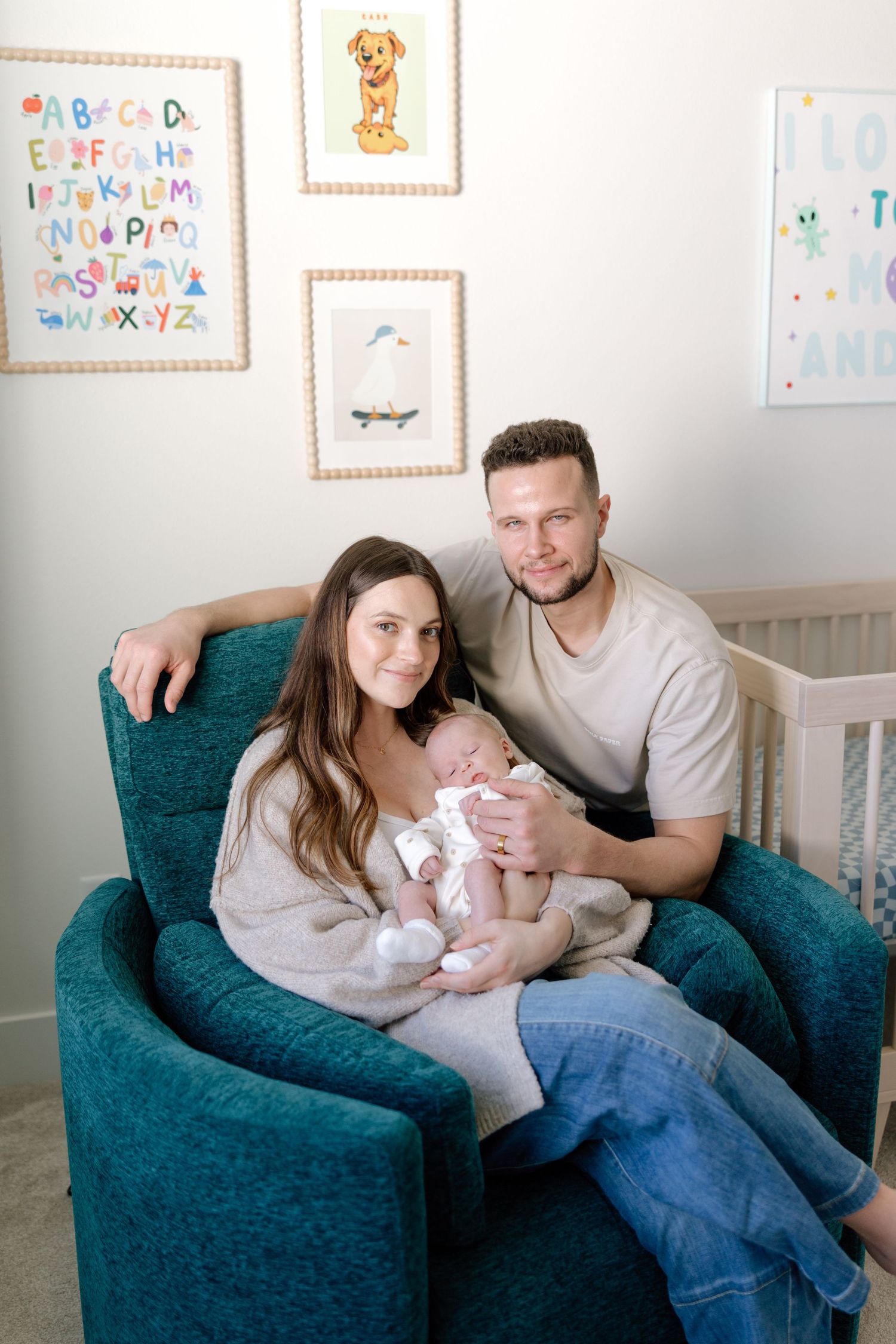 Happy new parents sit in a teal armchair, smiling warmly while holding their sleeping newborn in a decorated nursery.