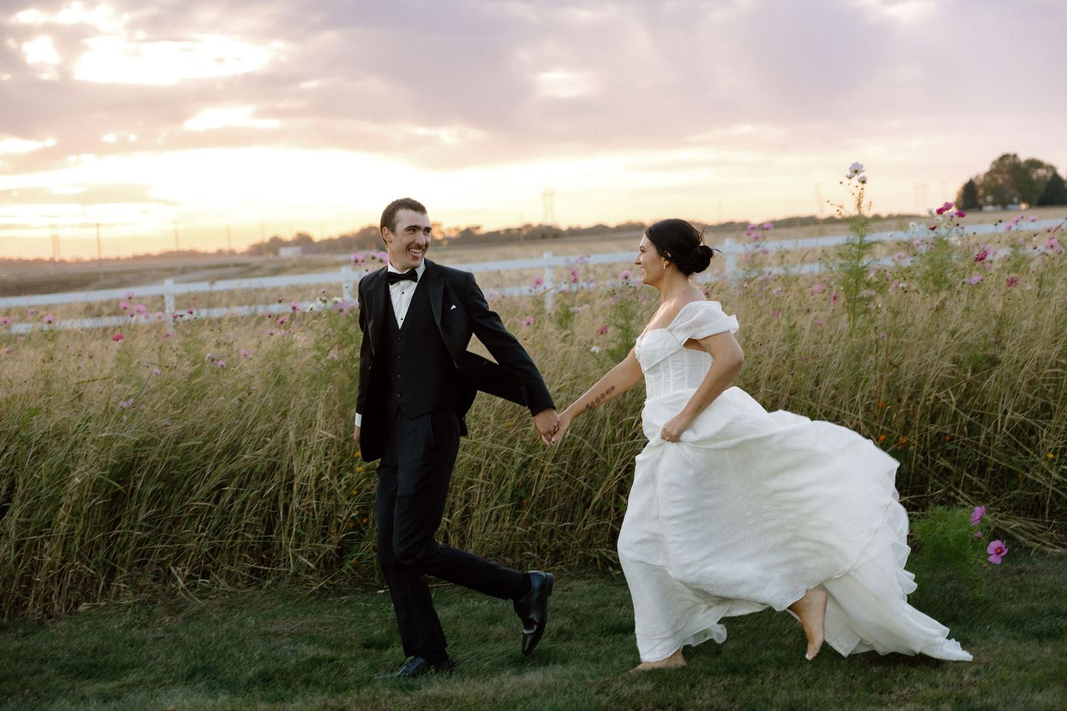 Bride and groom walk hand in hand through a lush green field at a stunning golden sunset.